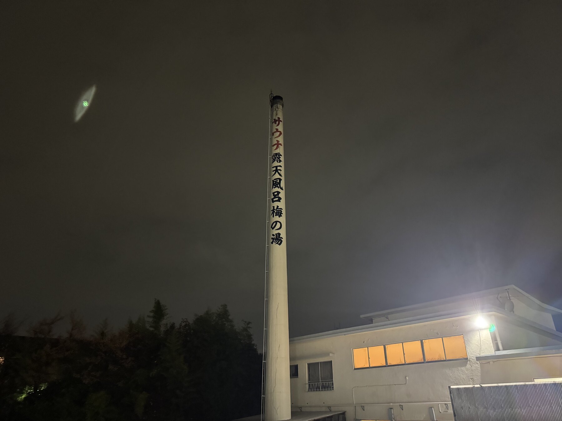 The tall illuminated chimney of Ume-no-Yu sentō at night, its vertical sign reading サウナ 露天風呂 梅の湯 (sauna, open-air bath, Ume-no-Yu), rising above the bathhouse roof.