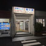 The lit night entrance of Ume-no-Yu sento in Chofu, Tokyo, with a blue sign reading Kenko Center Ume-no-Yu and a board listing its sauna, electric bath, herbal bath and open-air bath, beside a vending machine and shoe lockers.