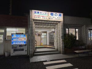 The lit night entrance of Ume-no-Yu sento in Chofu, Tokyo, with a blue sign reading Kenko Center Ume-no-Yu and a board listing its sauna, electric bath, herbal bath and open-air bath, beside a vending machine and shoe lockers.