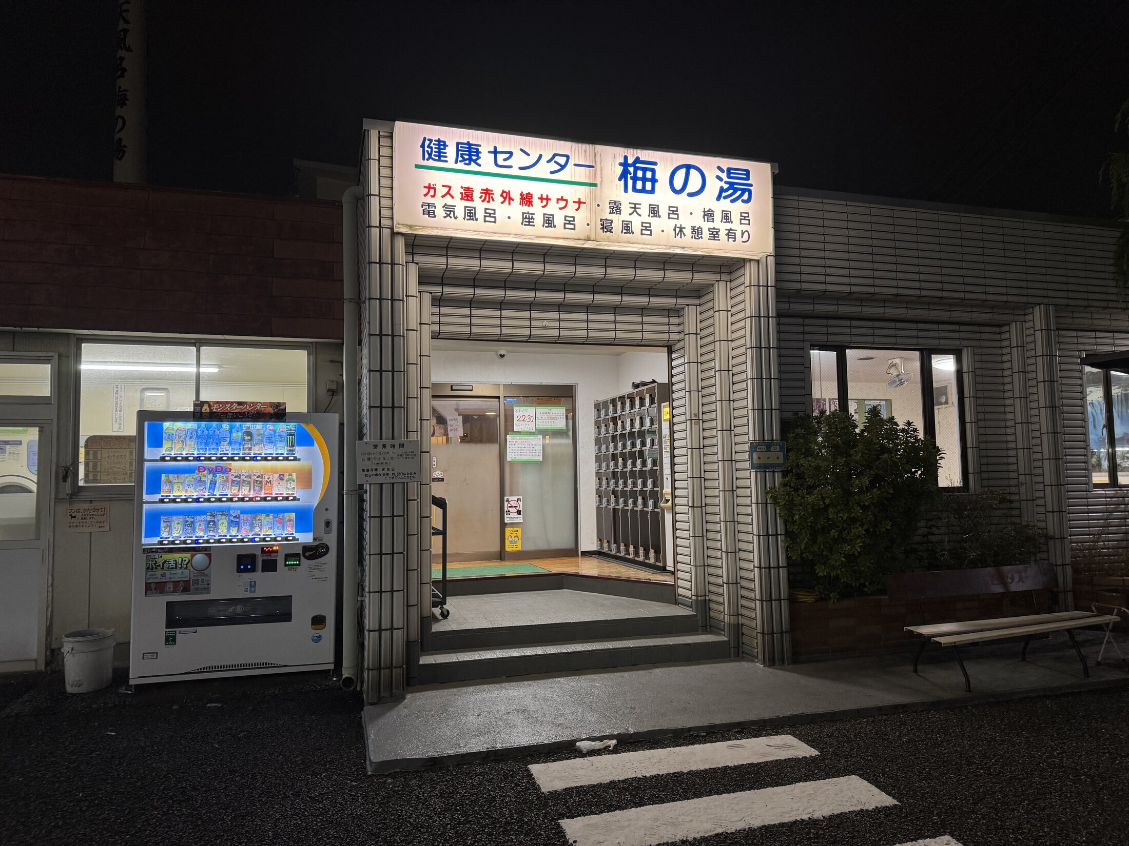 The lit night entrance of Ume-no-Yu sento in Chofu, Tokyo, with a blue sign reading Kenko Center Ume-no-Yu and a board listing its sauna, electric bath, herbal bath and open-air bath, beside a vending machine and shoe lockers.