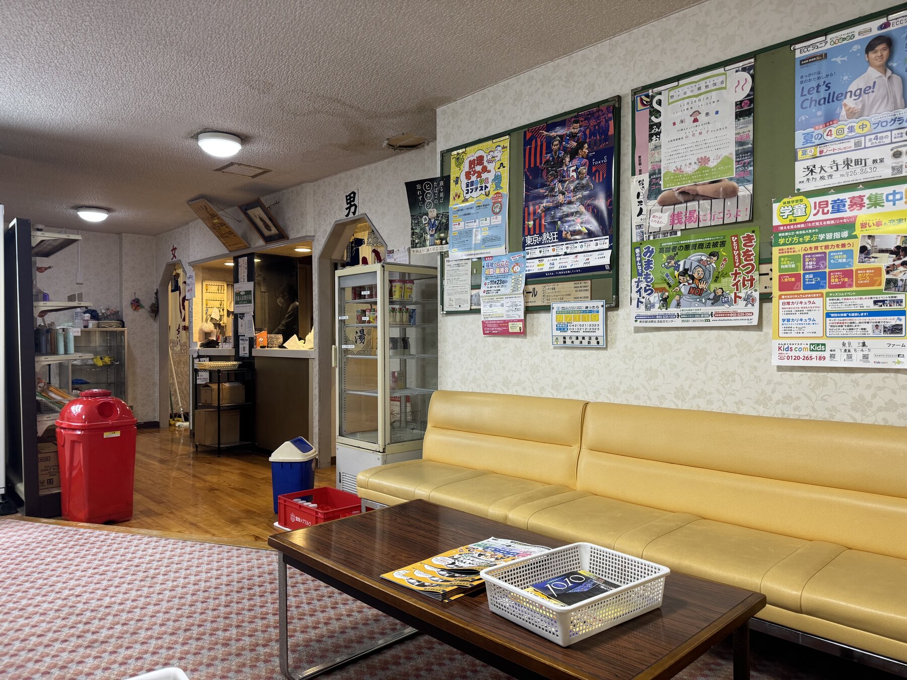 The retro lounge of Ume-no-Yu sentō with a yellow vinyl sofa, a drinks fridge, the men's (男) entrance curtain and community posters on the wall.