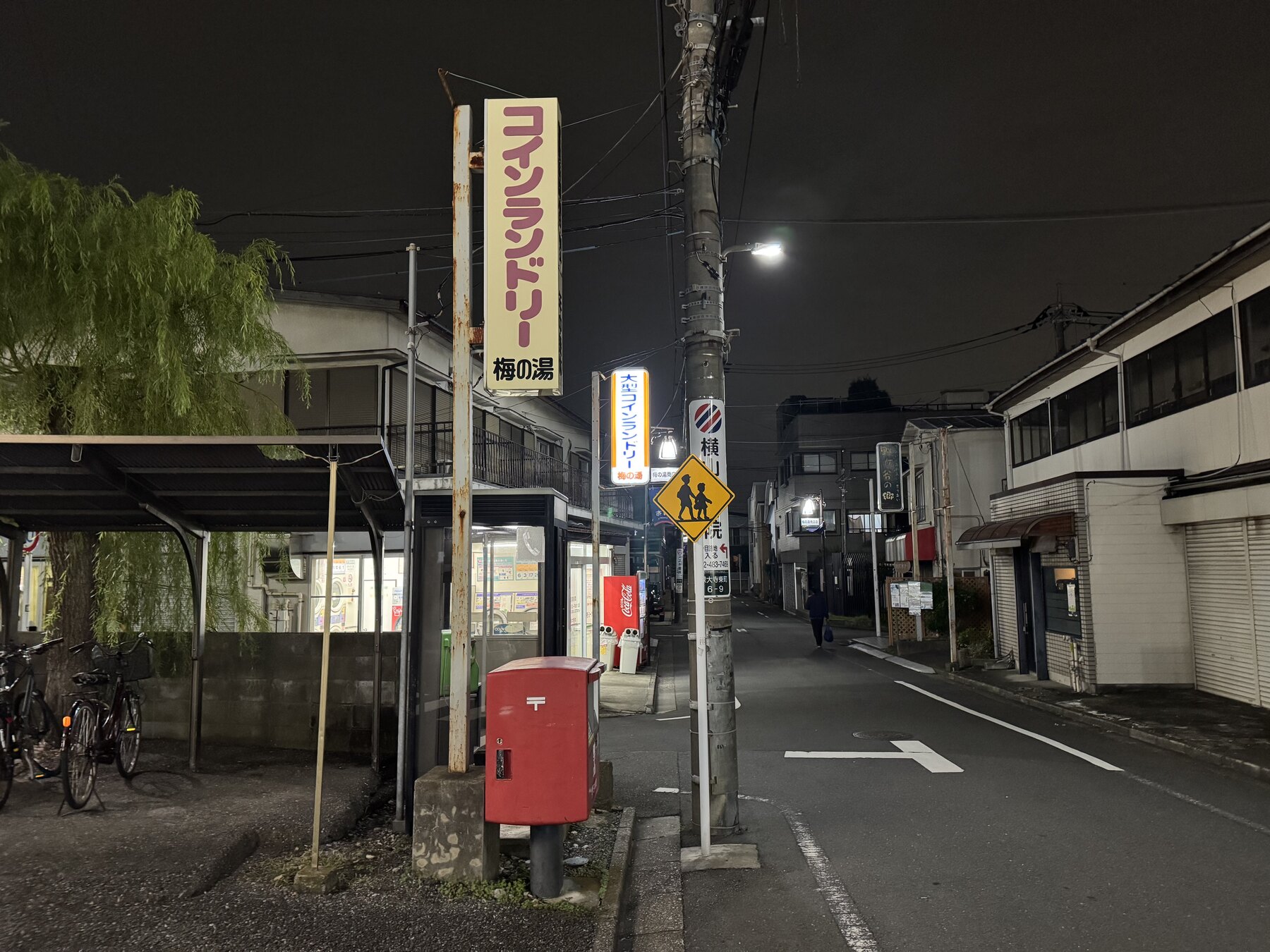 The quiet Chōfu street outside Ume-no-Yu at night, with the lit coin-laundry and Ume-no-Yu sign, a willow tree and a red postbox.