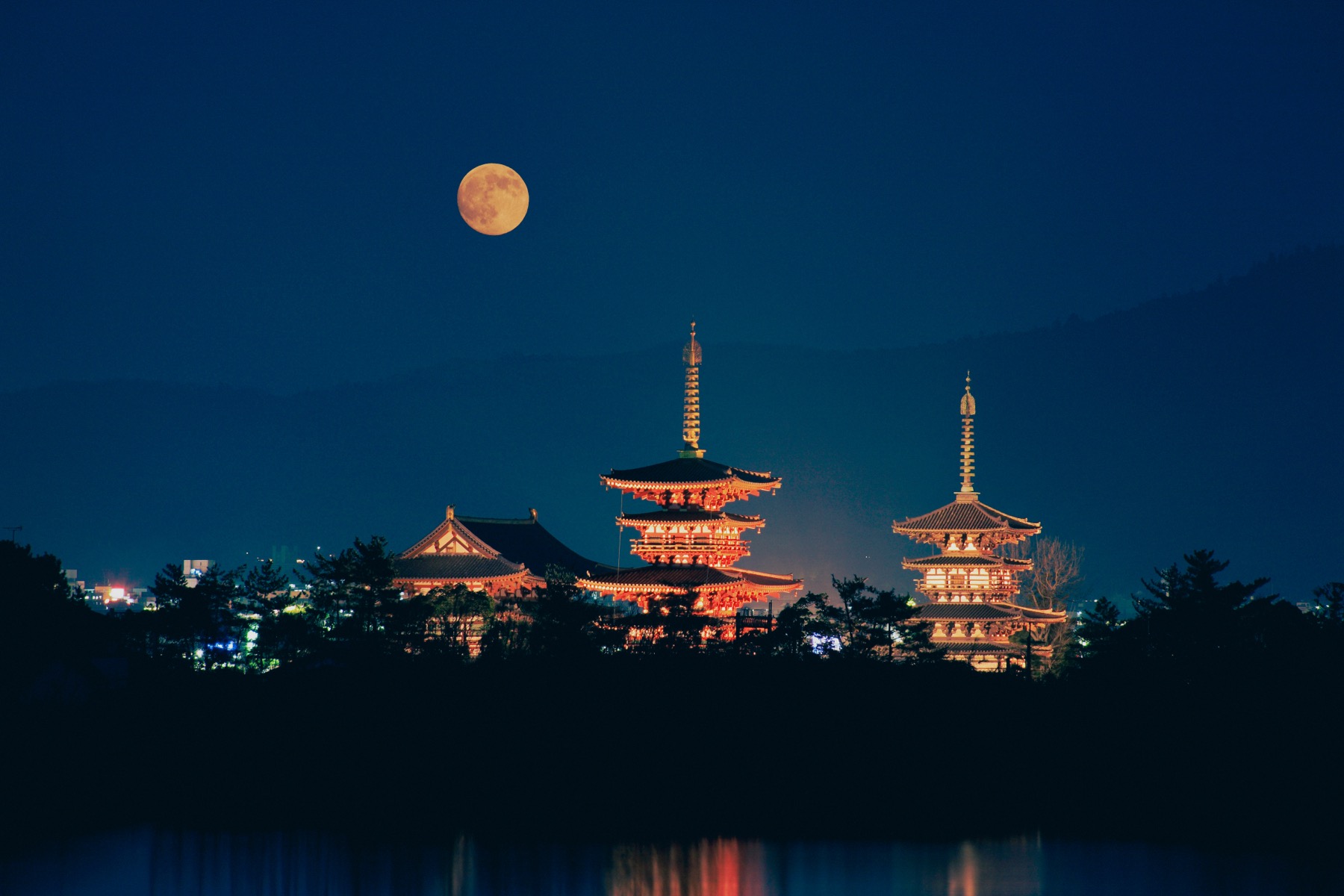 The two illuminated pagodas and Golden Hall of Yakushiji Temple in Nara glowing at night beneath a full moon, reflected in water with mountains behind, during an evening lighting.