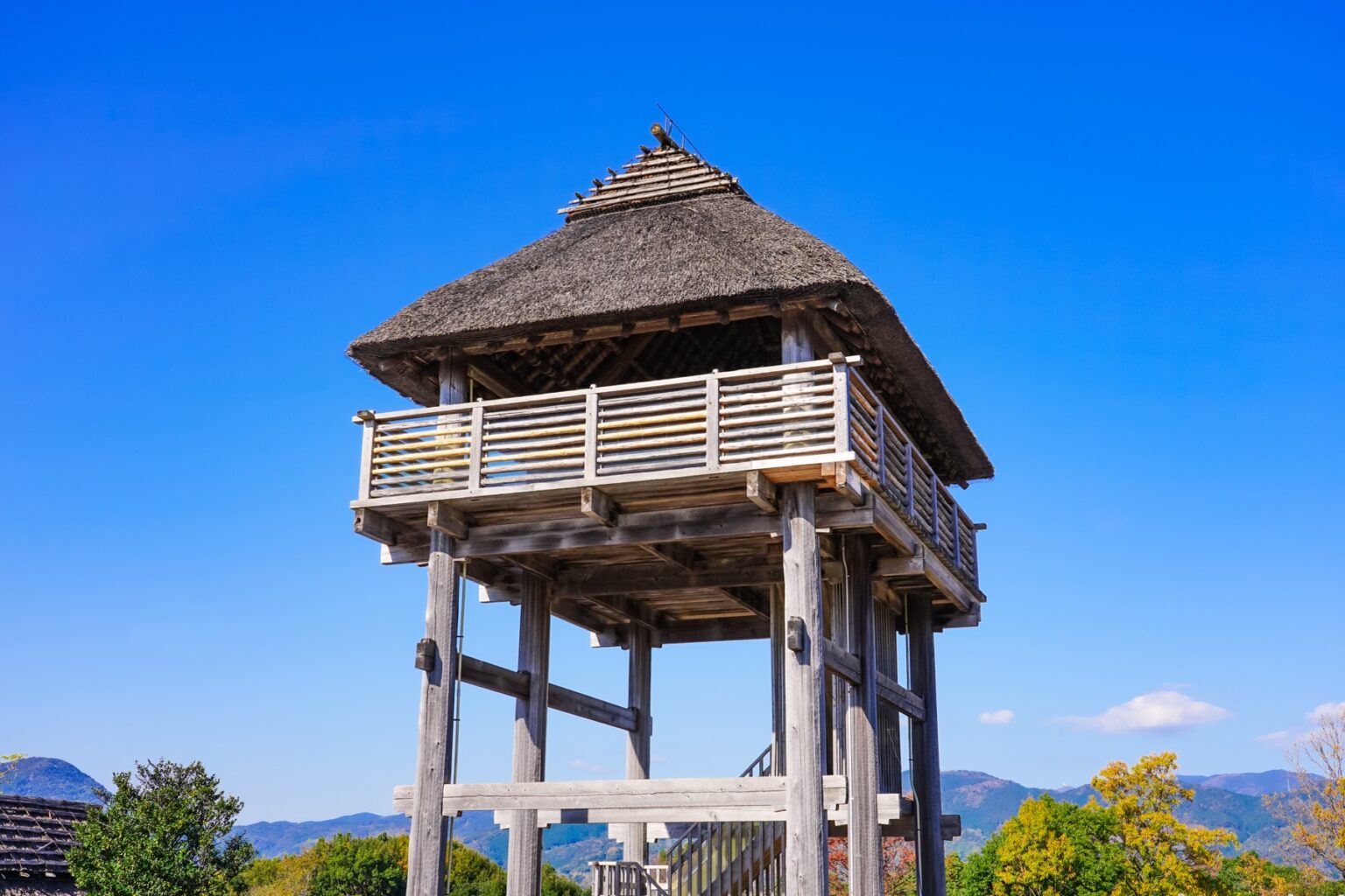 A reconstructed wooden watchtower with a thatched roof and open viewing platform at Yoshinogari Historical Park in Saga, a defensive lookout of the Yayoi-period moated settlement against a blue sky.