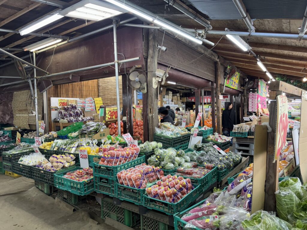 Open-air stalls at the Aozora Ichiba discount market near Lake Kawaguchi, Yamanashi, piled with crates of strawberries, mandarins, tomatoes and other fruit and vegetables under a covered roof.