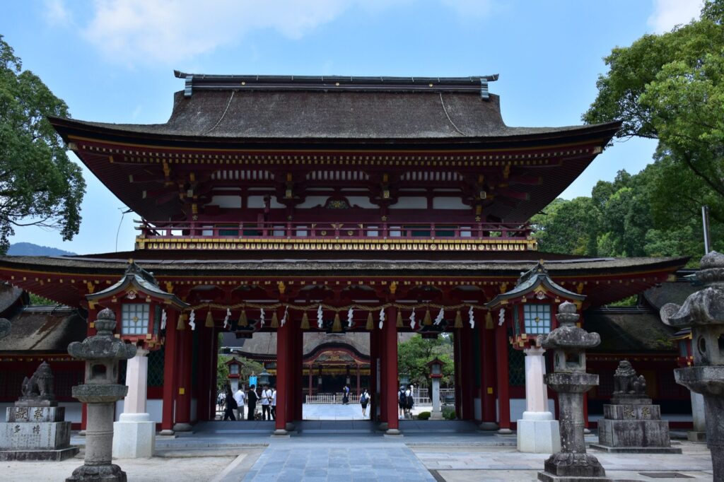 The vermilion two-storey romon gate of Dazaifu Tenmangu shrine framed by stone lanterns under a blue sky