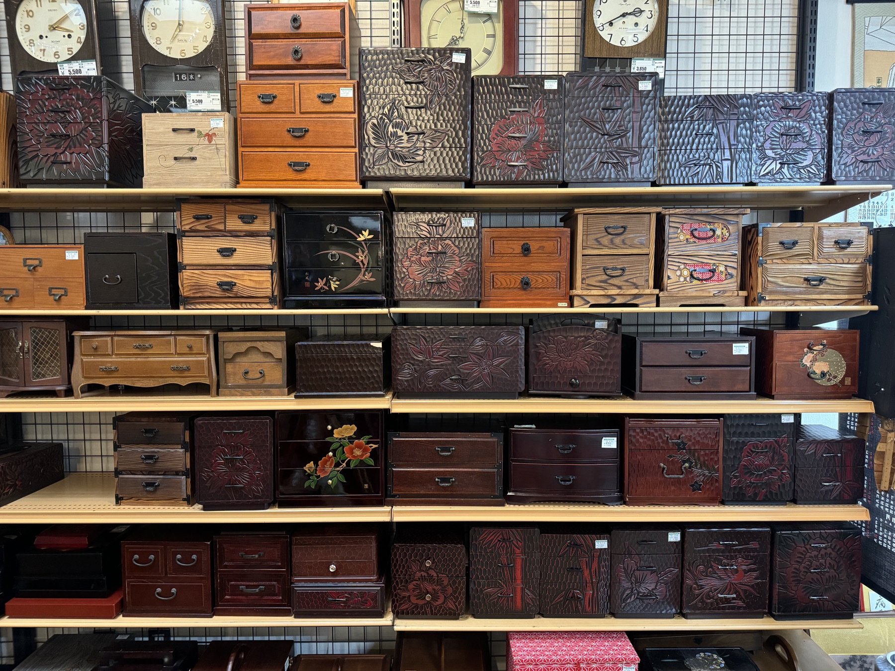 Shelves of carved wooden tansu chests, sewing boxes and small drawers with vintage wall clocks above them in the antique furniture section of Off House Hachioji Owada, Tokyo
