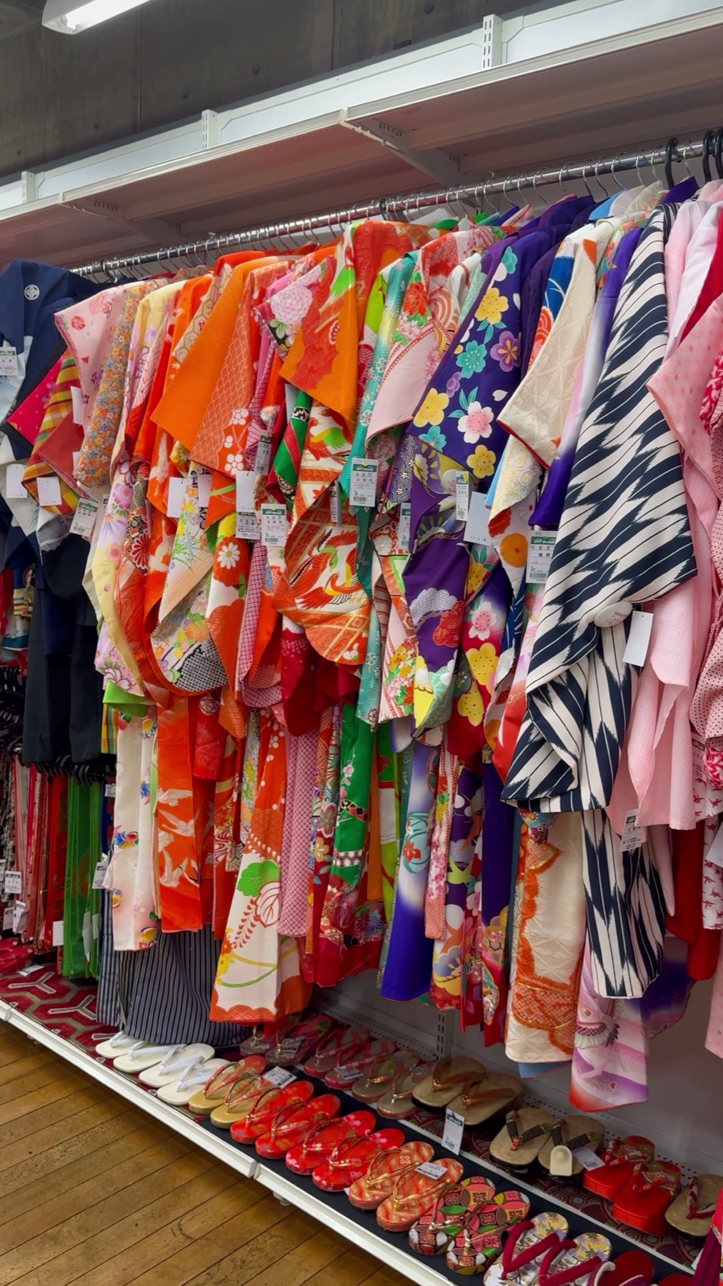 A rack of brightly coloured secondhand kimono and furisode above rows of woven zori and geta sandals on a patterned mat at the Off House store in Hachioji Owada, western Tokyo