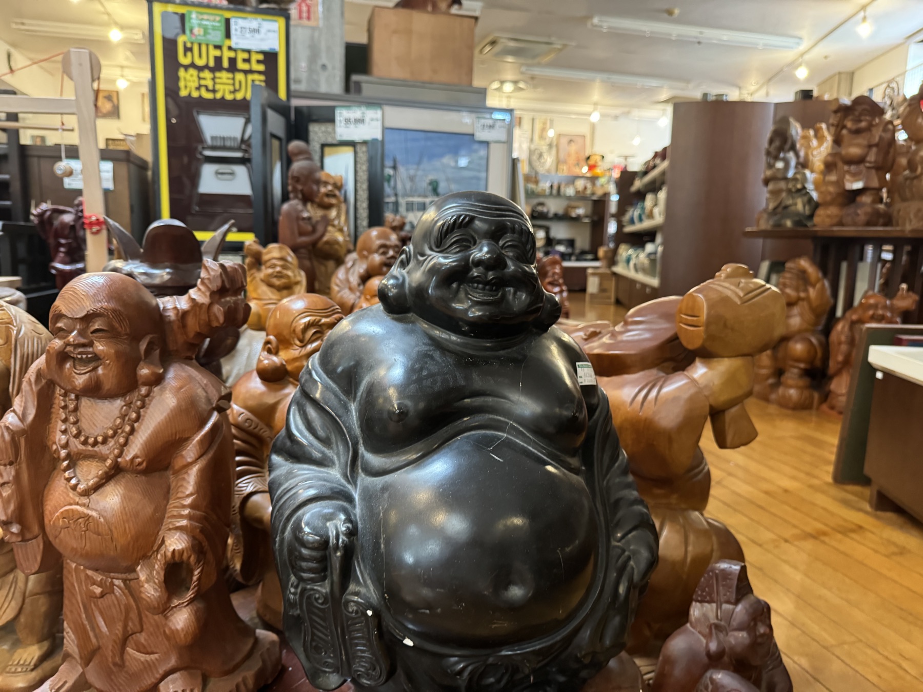 A large black laughing Hotei statue surrounded by carved wooden figures of lucky gods and laughing monks in the antiques area of the Hard Off Hachioji Owada reuse complex in Tokyo