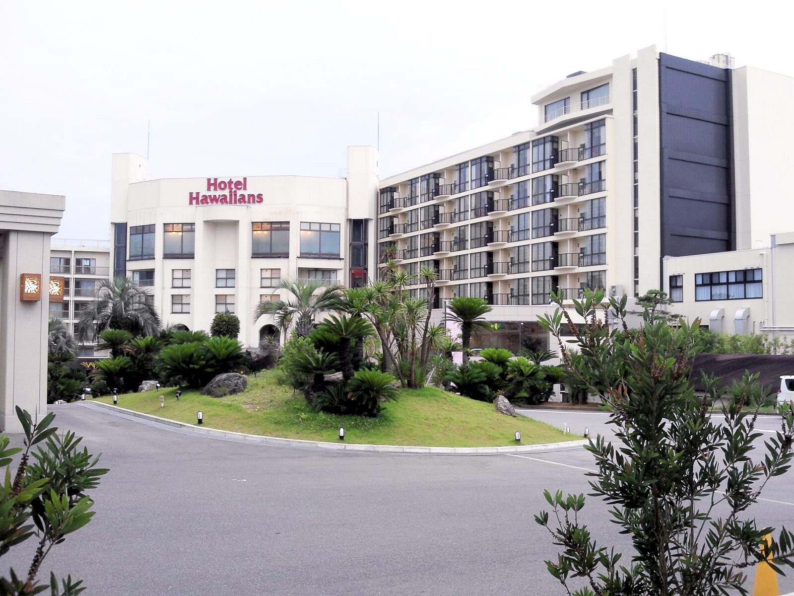 The exterior of Hotel Hawaiians, a white multi-storey hotel with the Hotel Hawaiians sign, palm trees and a landscaped driveway