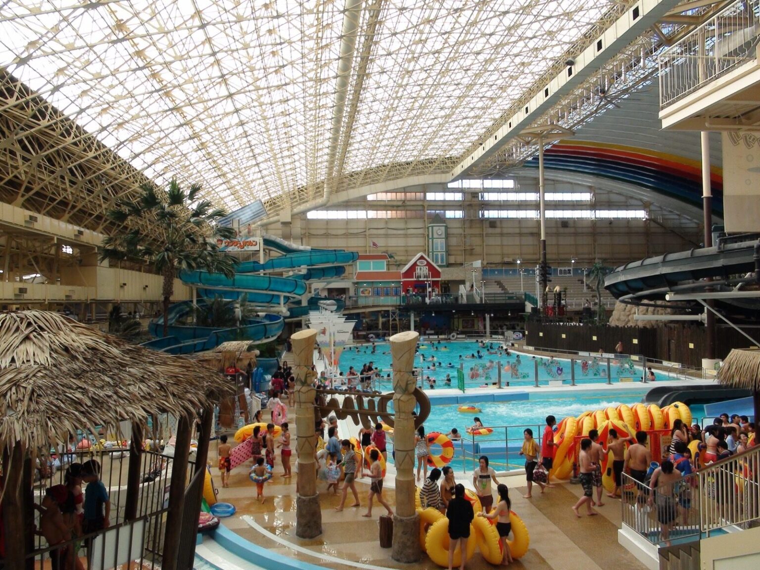 Crowds of visitors around the pools of the Spa Resort Hawaiians dome, with palm-thatched huts, yellow inflatable rings, water slides and a rainbow arch under the glass roof