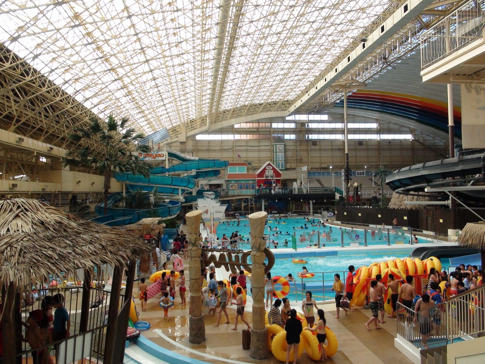 Crowds of visitors around the pools of the Spa Resort Hawaiians dome, with palm-thatched huts, yellow inflatable rings, water slides and a rainbow arch under the glass roof