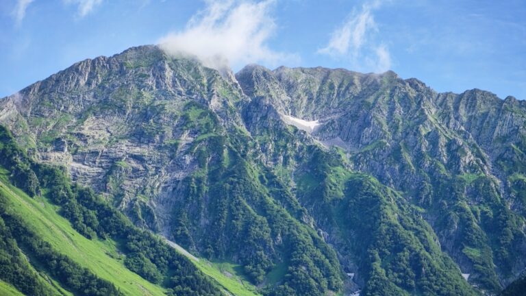 The jagged rock walls and green summer slopes of the Hotaka mountain range rising above Kamikochi in the Japanese Northern Alps, patches of late-season snow lingering in the high gullies under a clear blue July sky