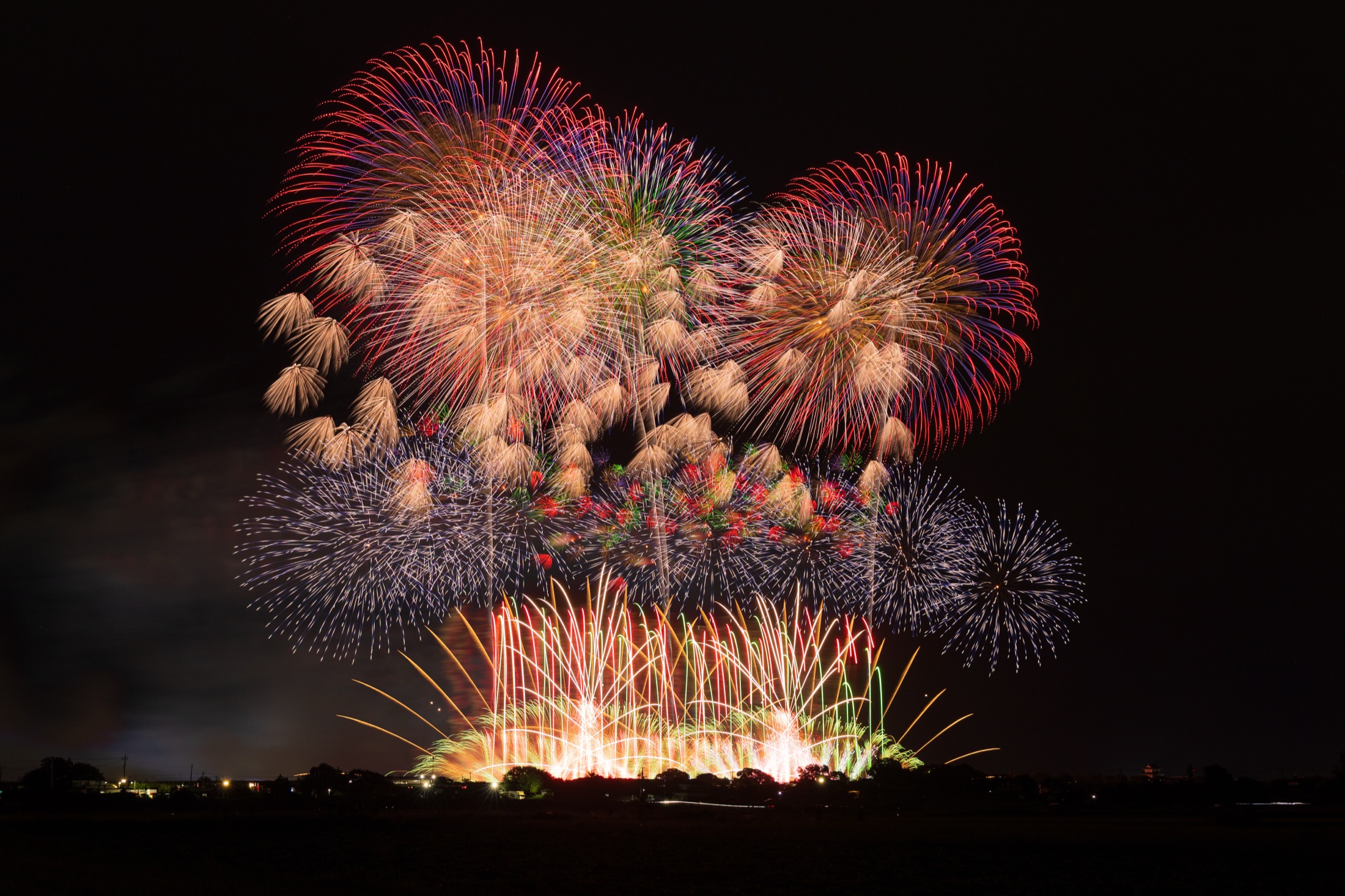 Enormous layered red and white chrysanthemum fireworks bursting in tiers above a dark river at a Japanese summer fireworks festival, golden comets fanning upward from the bank below the massive display