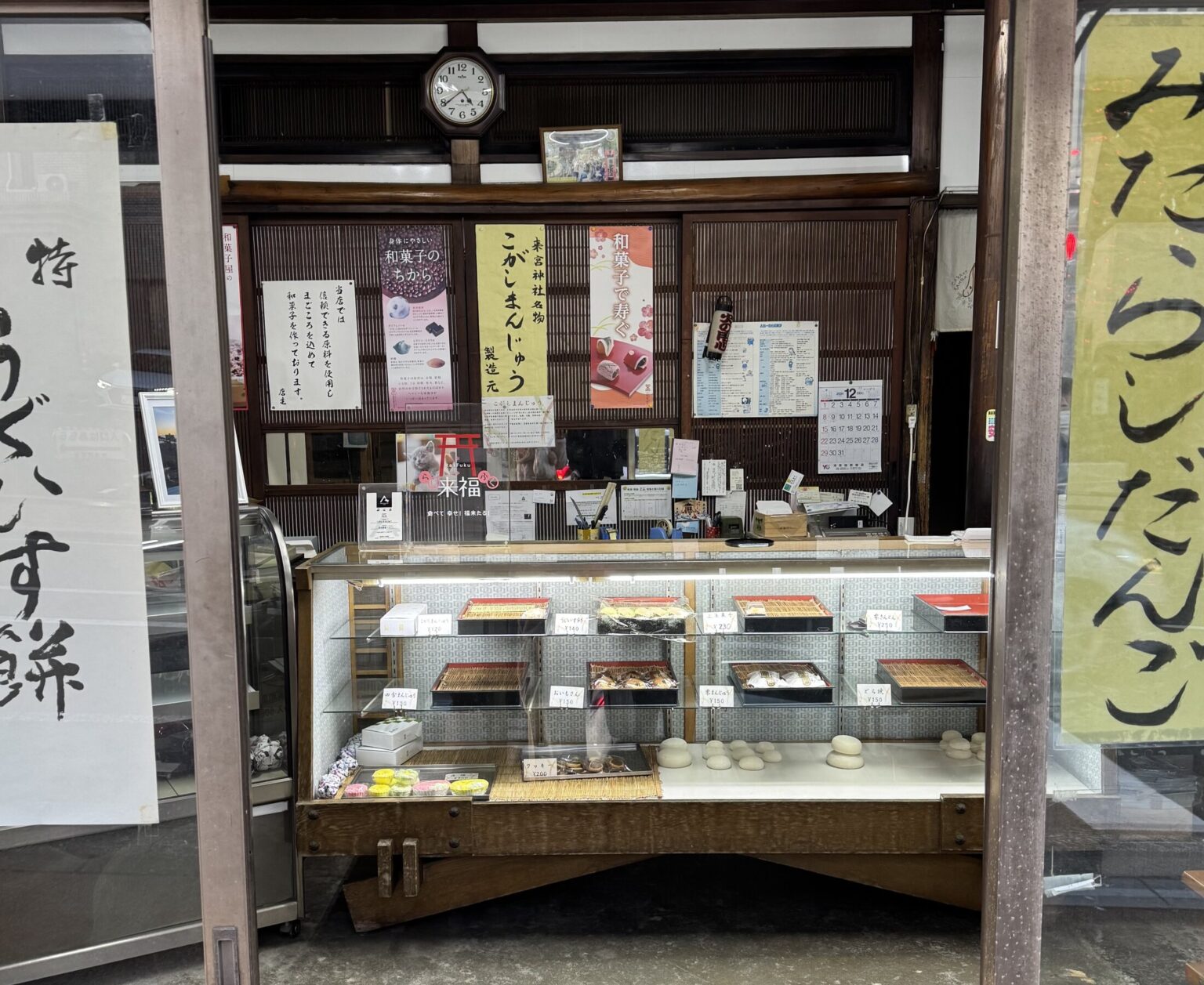 The old wood-framed display case of Kinomiya Kenko Pan in Atami, Shizuoka, under a banner reading Kinomiya Shrine specialty kogashi manju, with trays of Japanese sweets priced from about 120 to 290 yen.