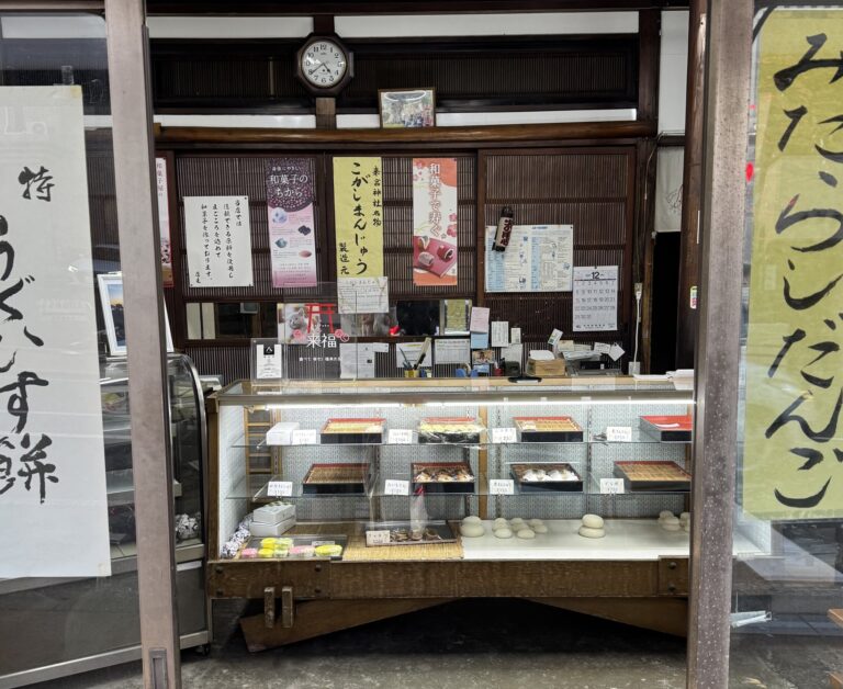 The old wood-framed display case of Kinomiya Kenko Pan in Atami, Shizuoka, under a banner reading Kinomiya Shrine specialty kogashi manju, with trays of Japanese sweets priced from about 120 to 290 yen.
