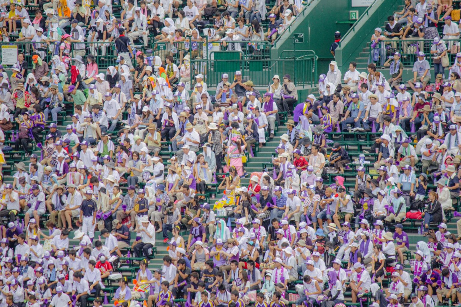 A packed Alps stand at Hanshin Koshien Stadium during the summer high school baseball tournament, thousands of spectators under a hazy summer sky