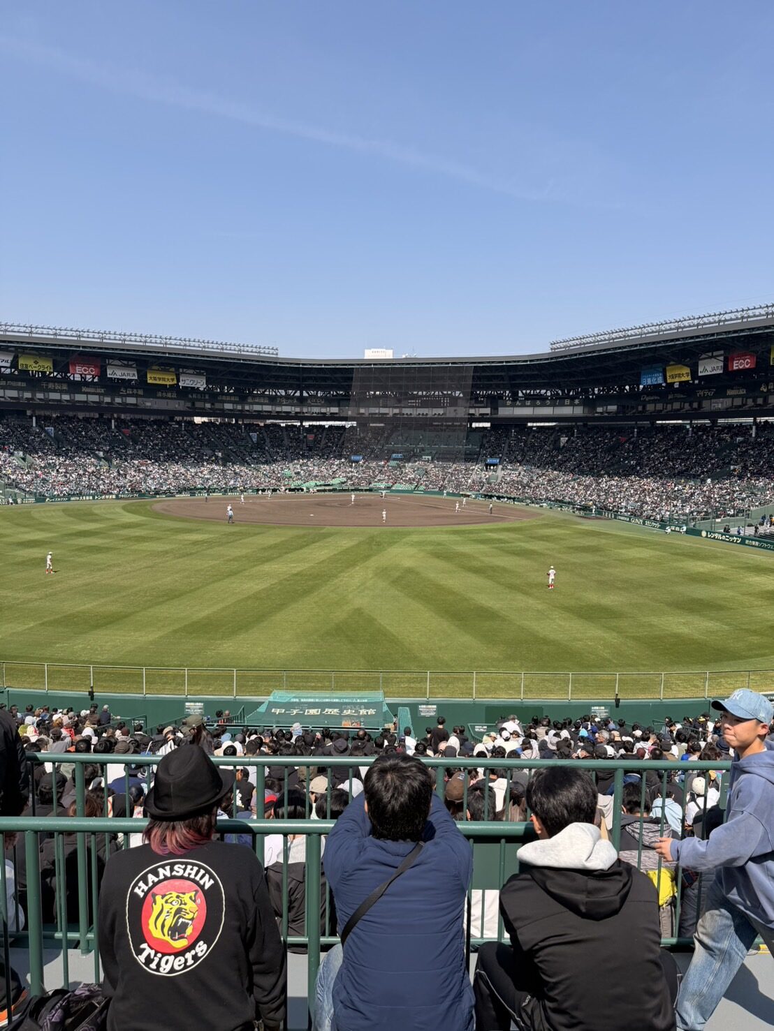Spectators at the outfield fence of Hanshin Koshien Stadium watching play, one fan in a Hanshin Tigers jacket, the Koshien History Museum banner visible on the outfield wall
