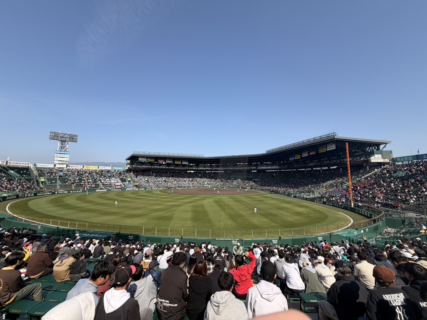 A wide view across the outfield grass of Hanshin Koshien Stadium from the packed right-field stands, the covered grandstand curving around the diamond under a clear blue sky