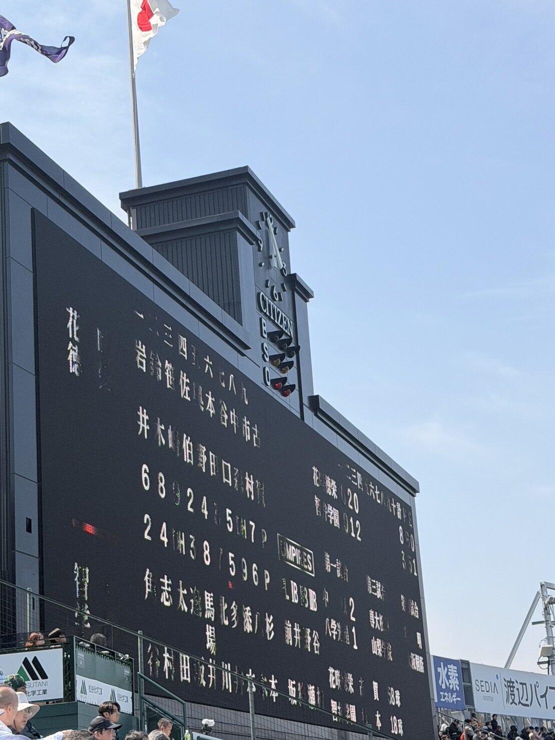 The famous black main scoreboard of Hanshin Koshien Stadium with its CITIZEN clock tower, player names in white Japanese characters and the national flag flying above