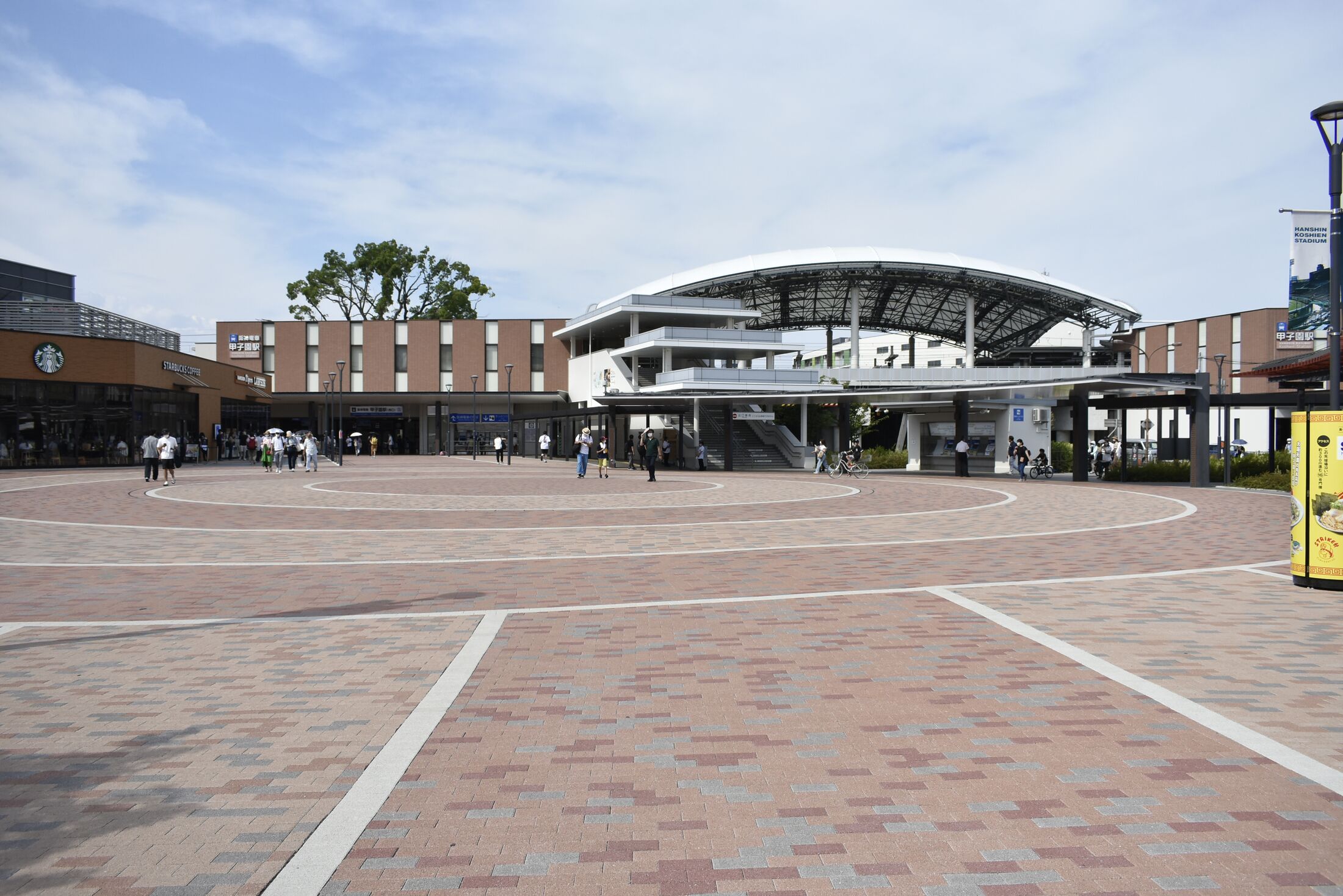 The curved white roof of Hanshin Koshien Station across its wide brick forecourt on a summer day, with the Corowa Koshien shopping centre to the left