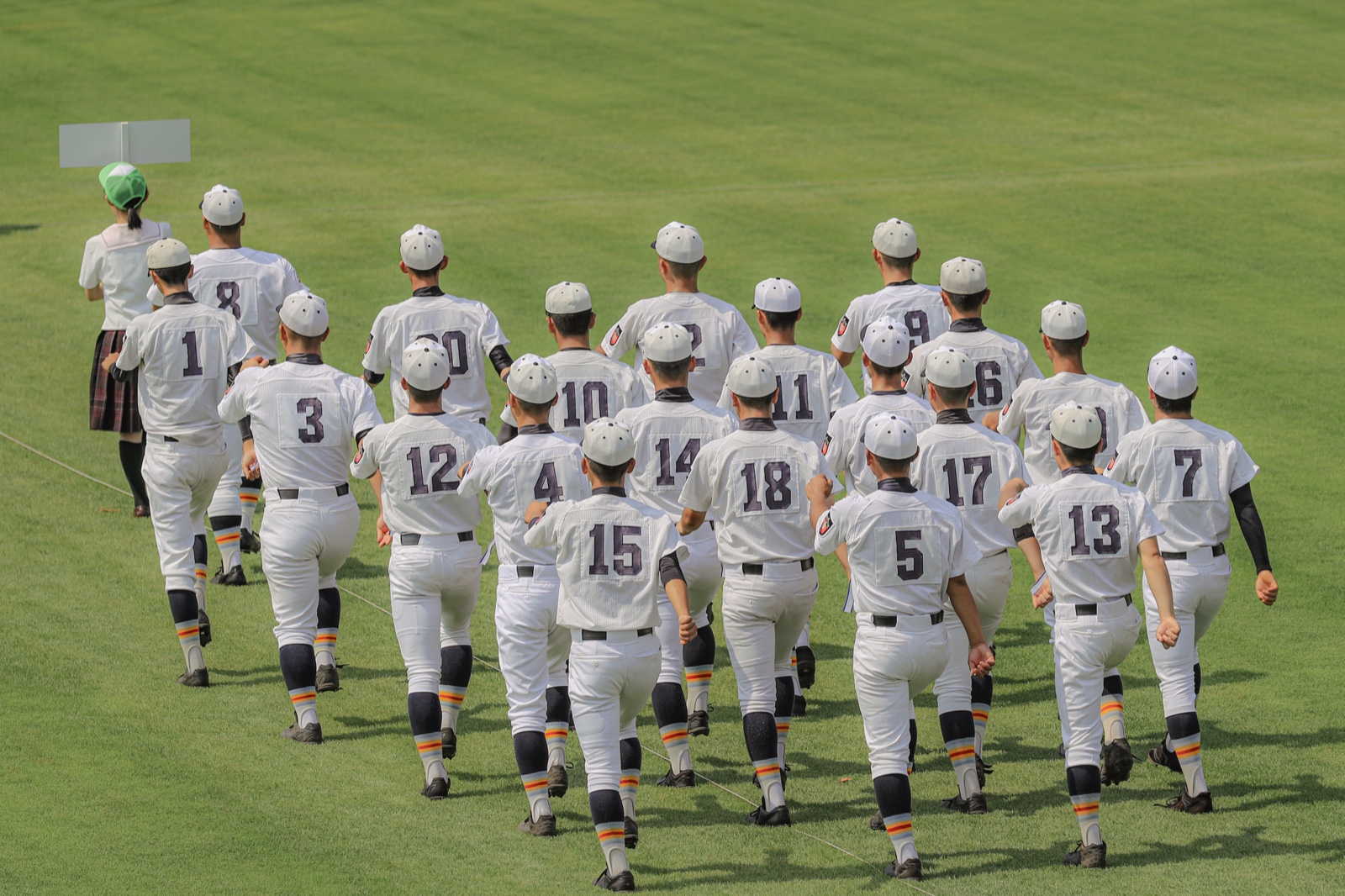 A high school baseball team jogging onto the field in numbered uniforms at Koshien