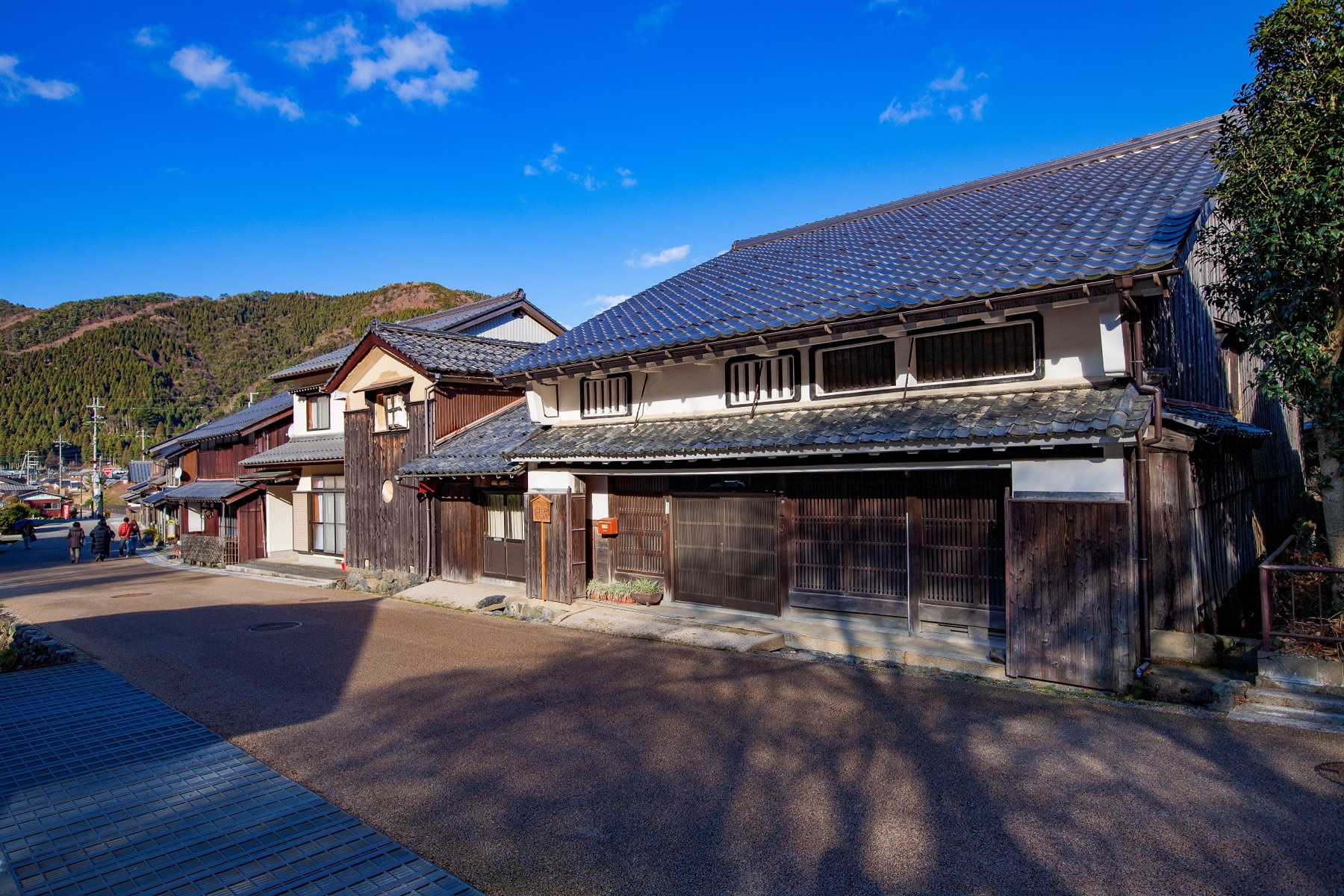 A large two-storey machiya with white plaster dark wood and a tiled roof on the street of Kumagawa-juku with forested mountains rising behind