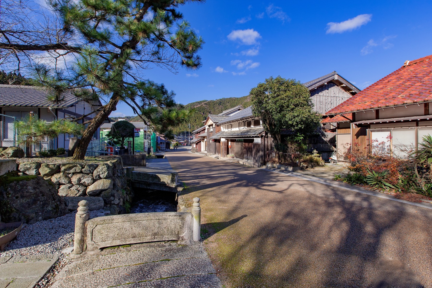 A small stone bridge over a water channel with a pine tree and traditional tile-roofed houses along the street of Kumagawa-juku Fukui and mountains behind