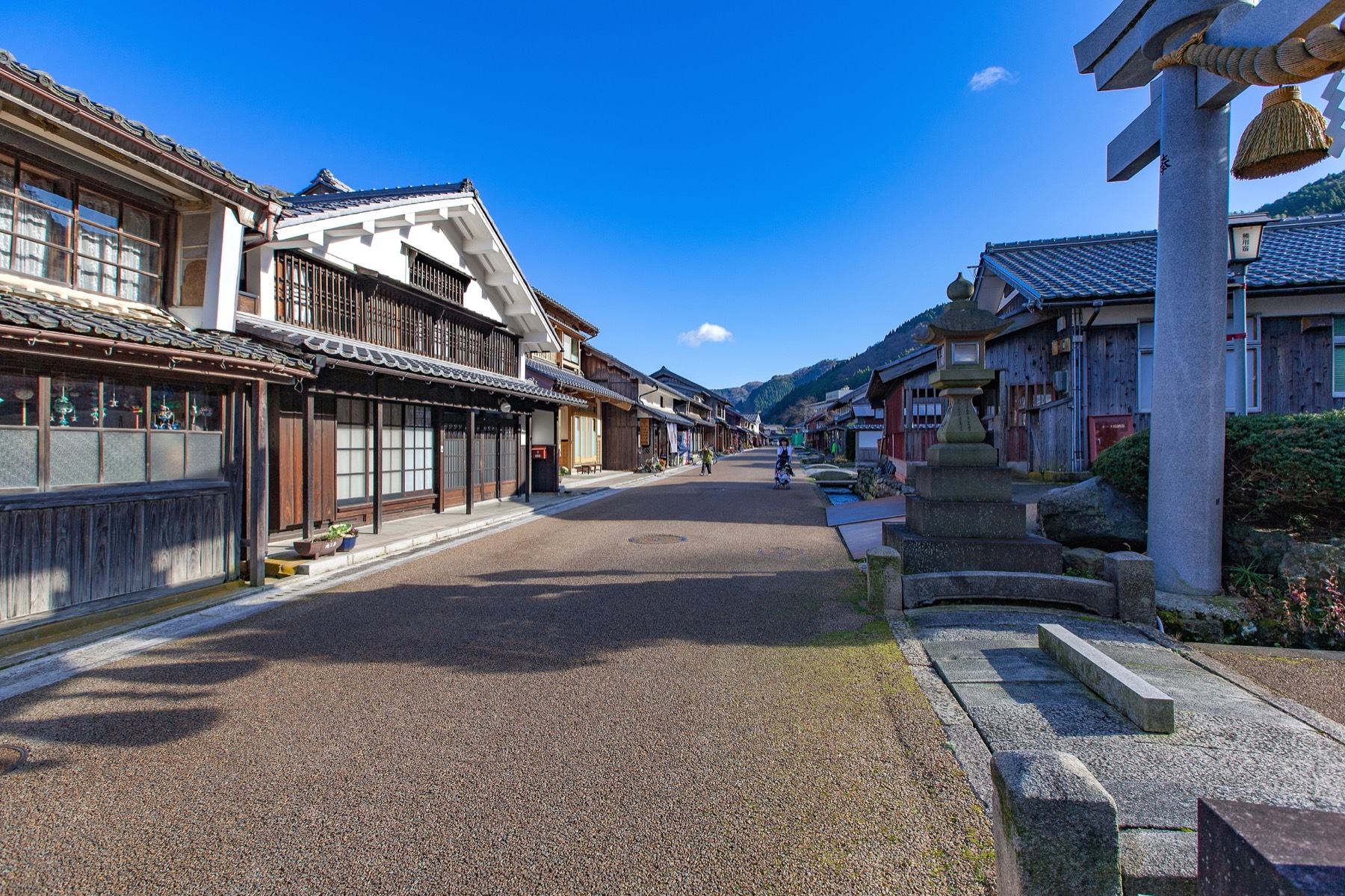 A stone torii gate and lantern beside the machiya-lined street of Kumagawa-juku post town under a clear blue sky with mountains at the end