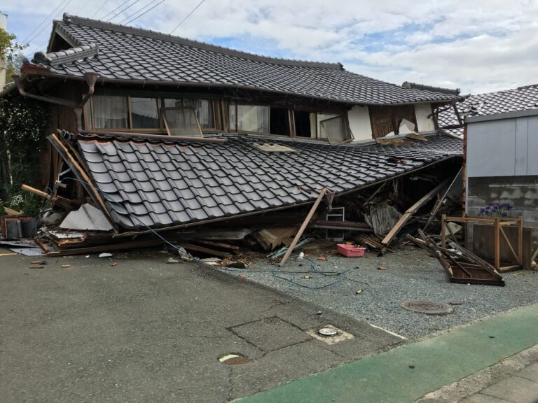 A single-storey Japanese house with a heavy tiled roof fully collapsed onto its ground floor after the April 2016 Kumamoto earthquake, timber and roof tiles spilling across the pavement in front