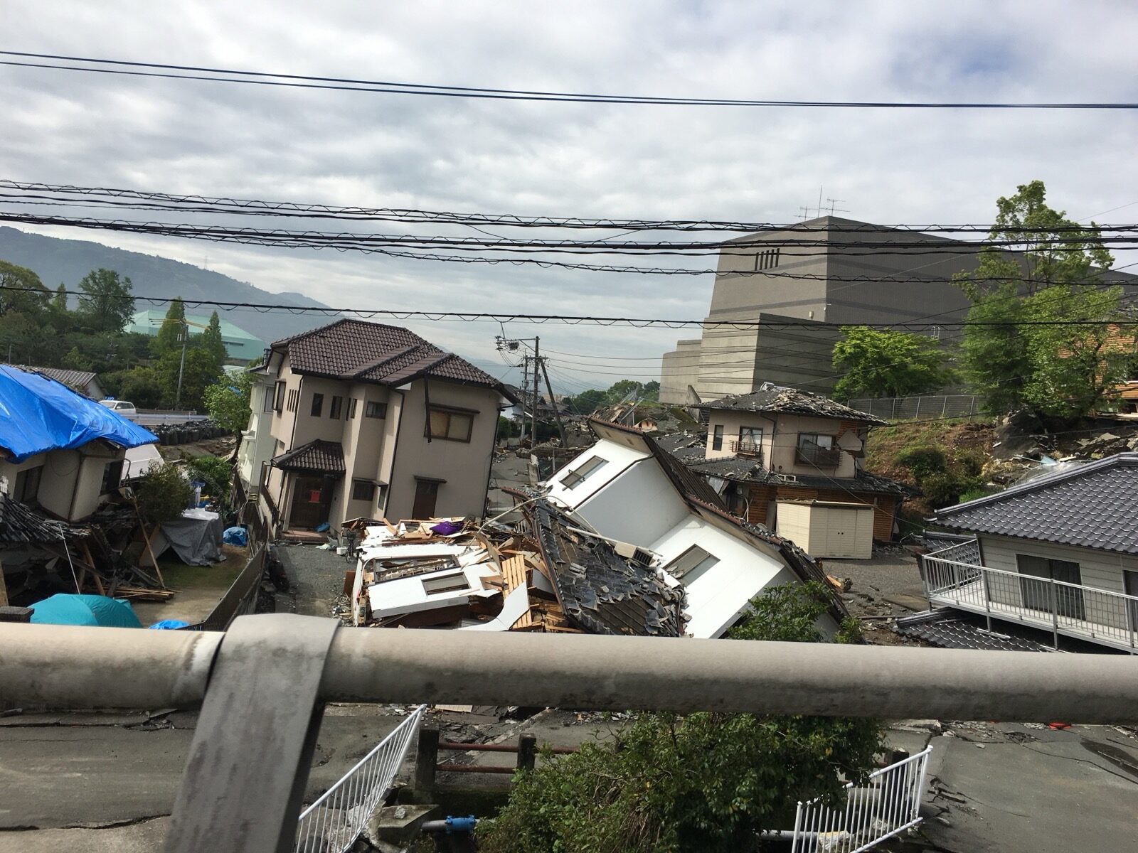 A hillside of houses leaning, collapsed and covered with blue tarpaulins after the April 2016 Kumamoto earthquake, seen from a road bridge
