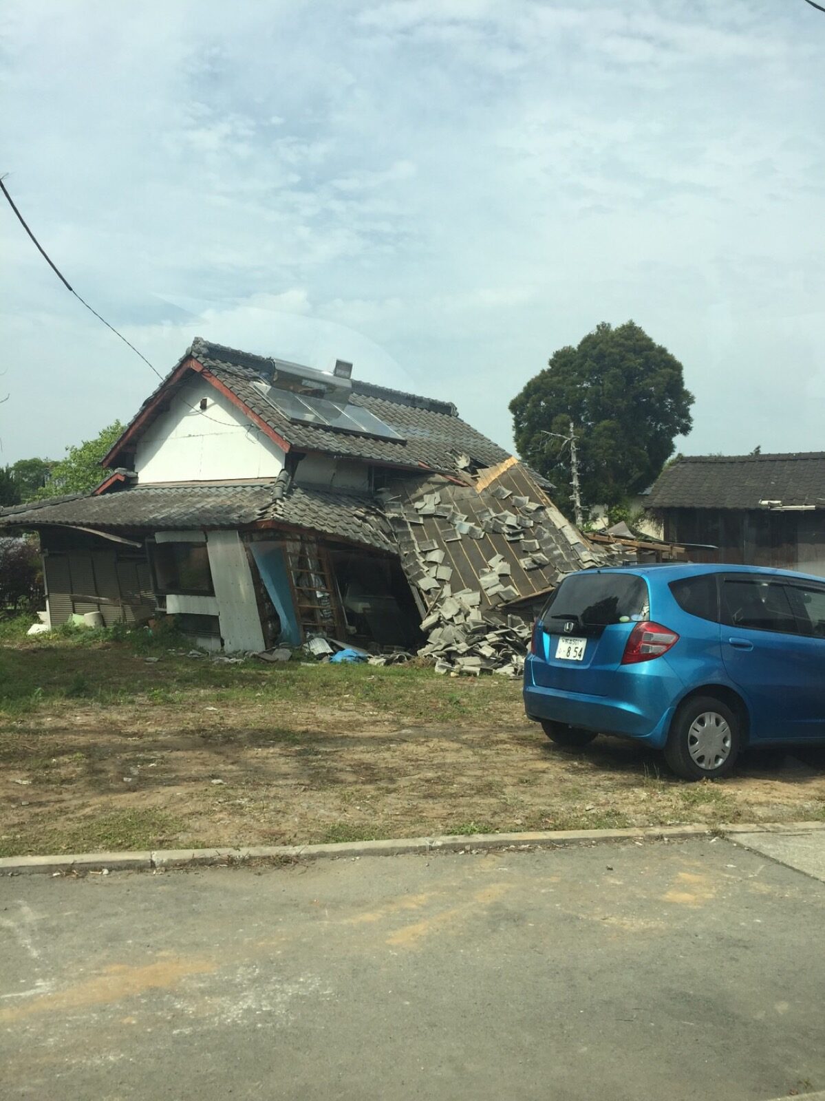 A single-storey house crushed and leaning with its wall fallen away next to a small blue car after the April 2016 Kumamoto earthquake