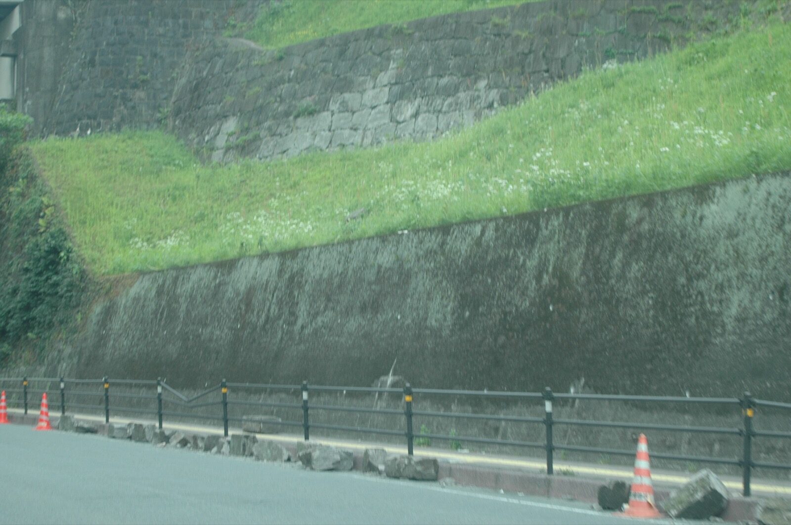 Rocks and soil fallen from a steep slope onto a road, partly blocked by a traffic cone, with a van passing, after the April 2016 Kumamoto earthquake