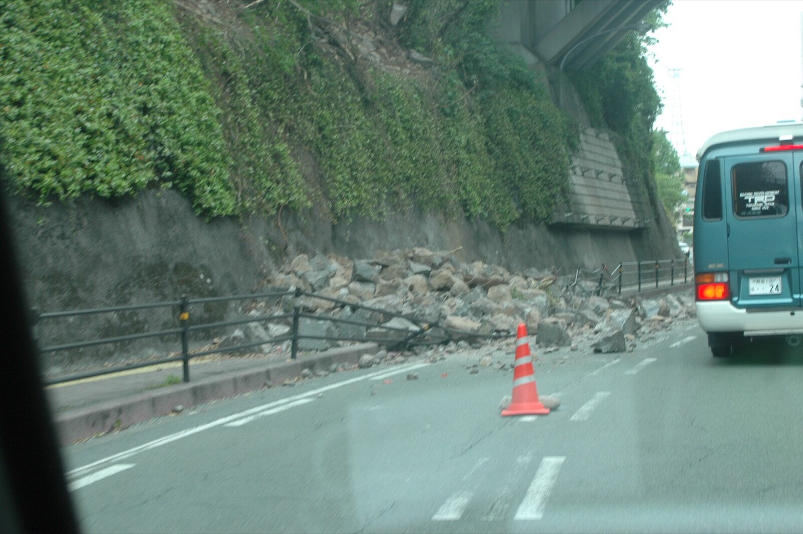 A blue building awning and facade panels torn loose and crashed onto a downtown pavement and pedestrian crossing after the April 2016 Kumamoto earthquake, with people inspecting the debris