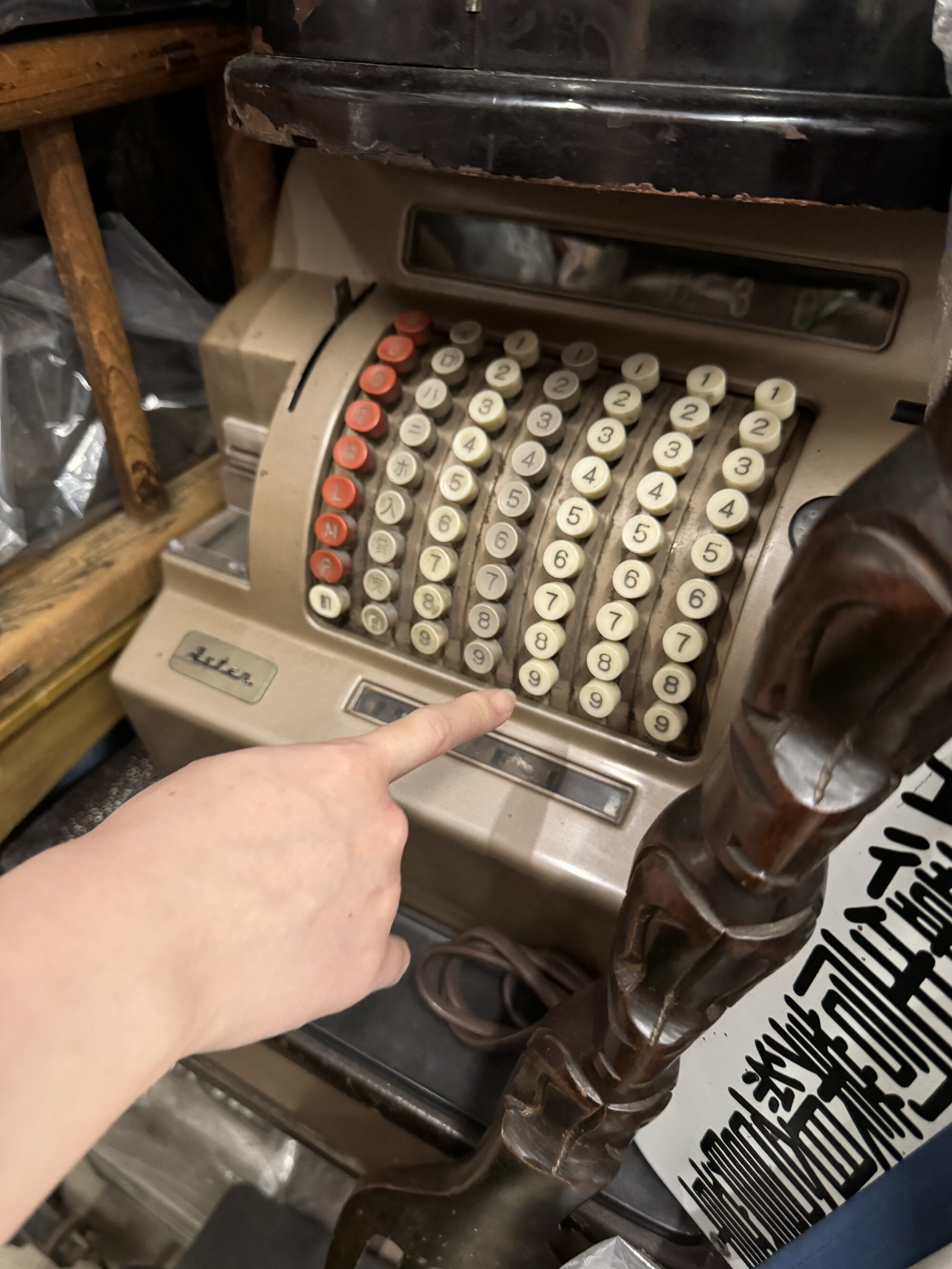 A hand pointing at an antique brass manual cash register with rows of number keys, one of the vintage finds among the goods at a local Kyoto recycling shop.