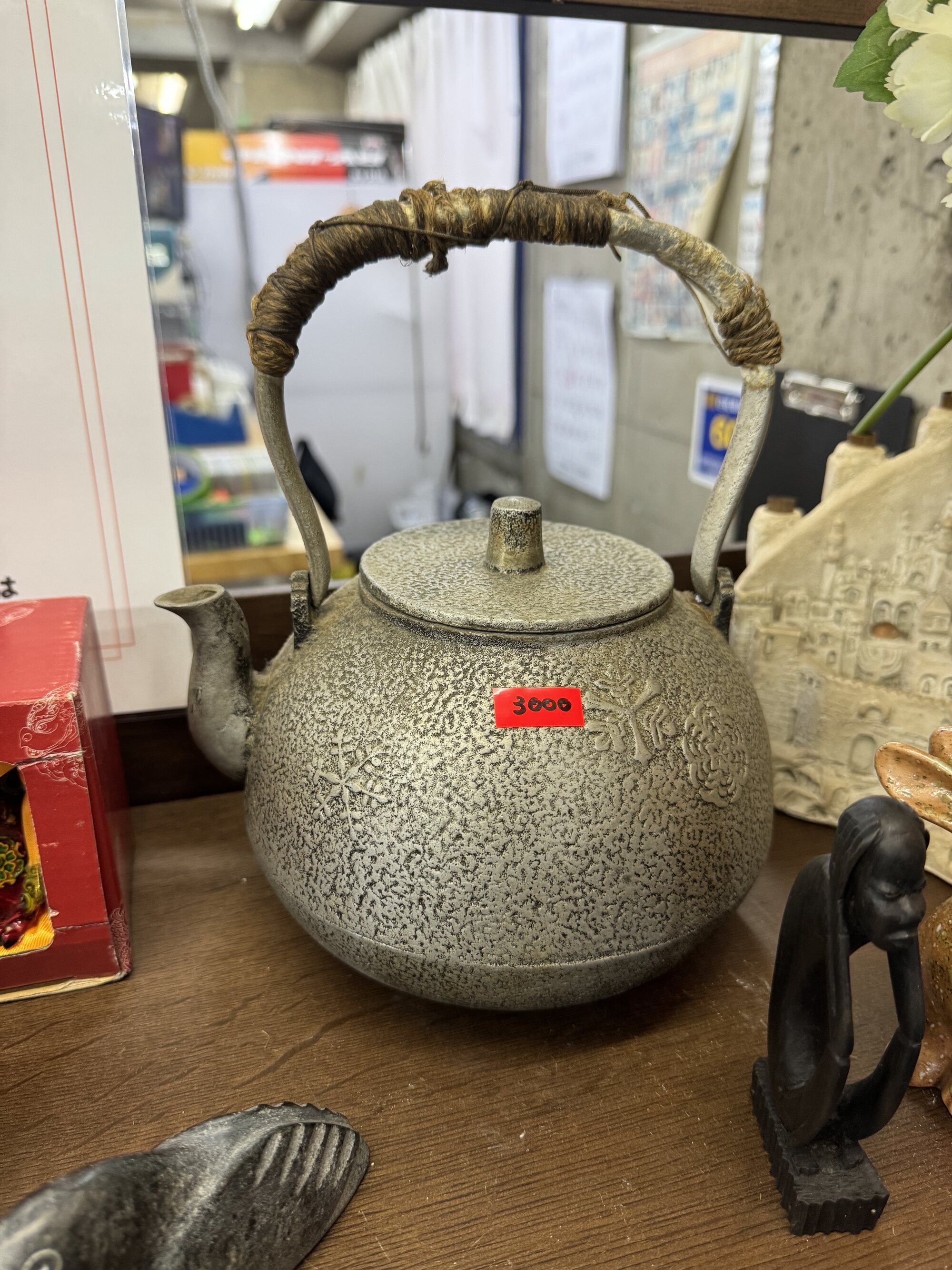 An antique cast-iron tetsubin kettle with a woven bamboo handle and a price tag, set on a wooden table among other used goods at a local Kyoto recycling shop.