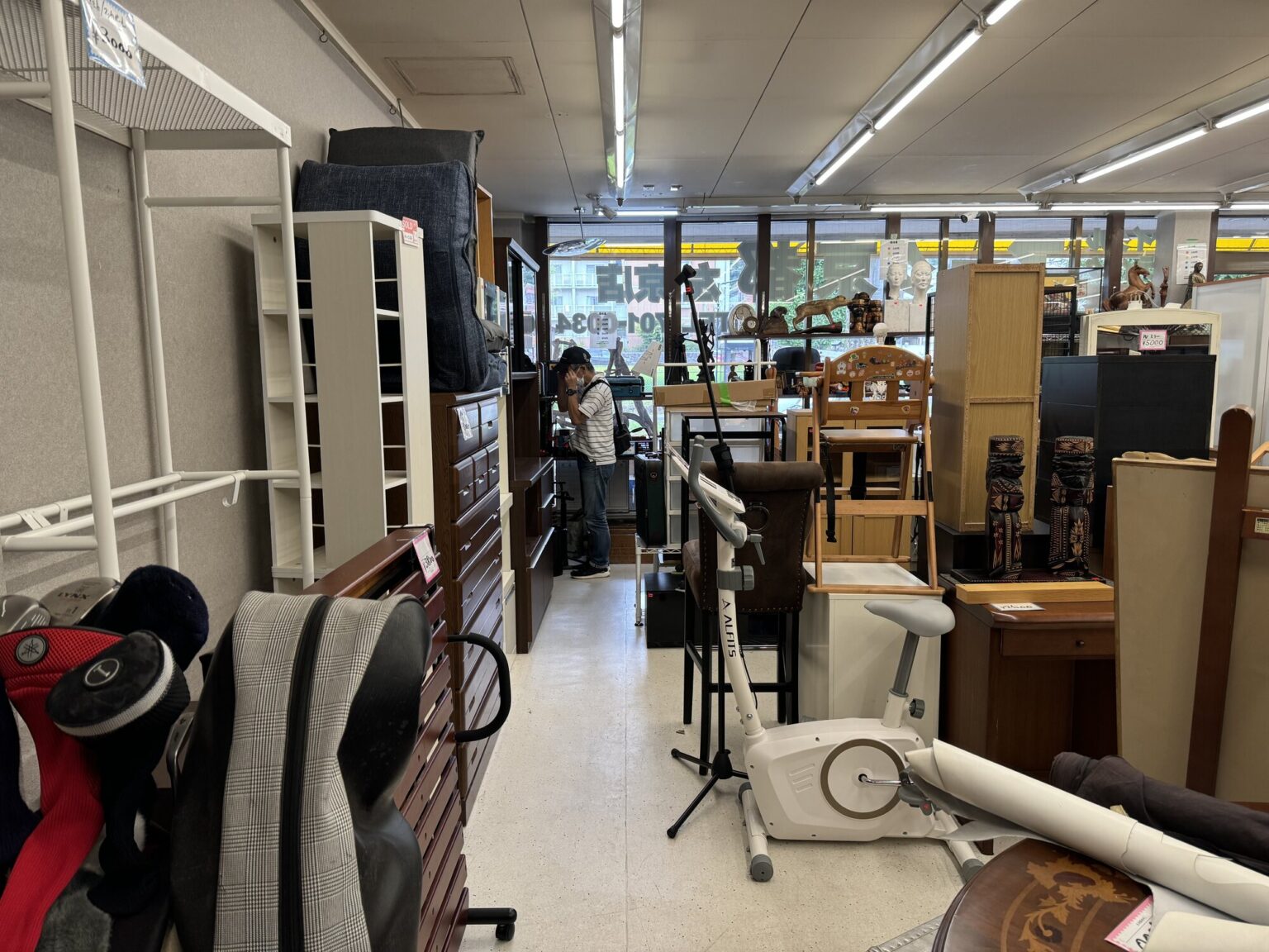 A packed aisle inside a local Kyoto recycling shop, crammed with used furniture, chests of drawers, chairs, golf clubs, an exercise bike and carved wooden folk items under fluorescent light.