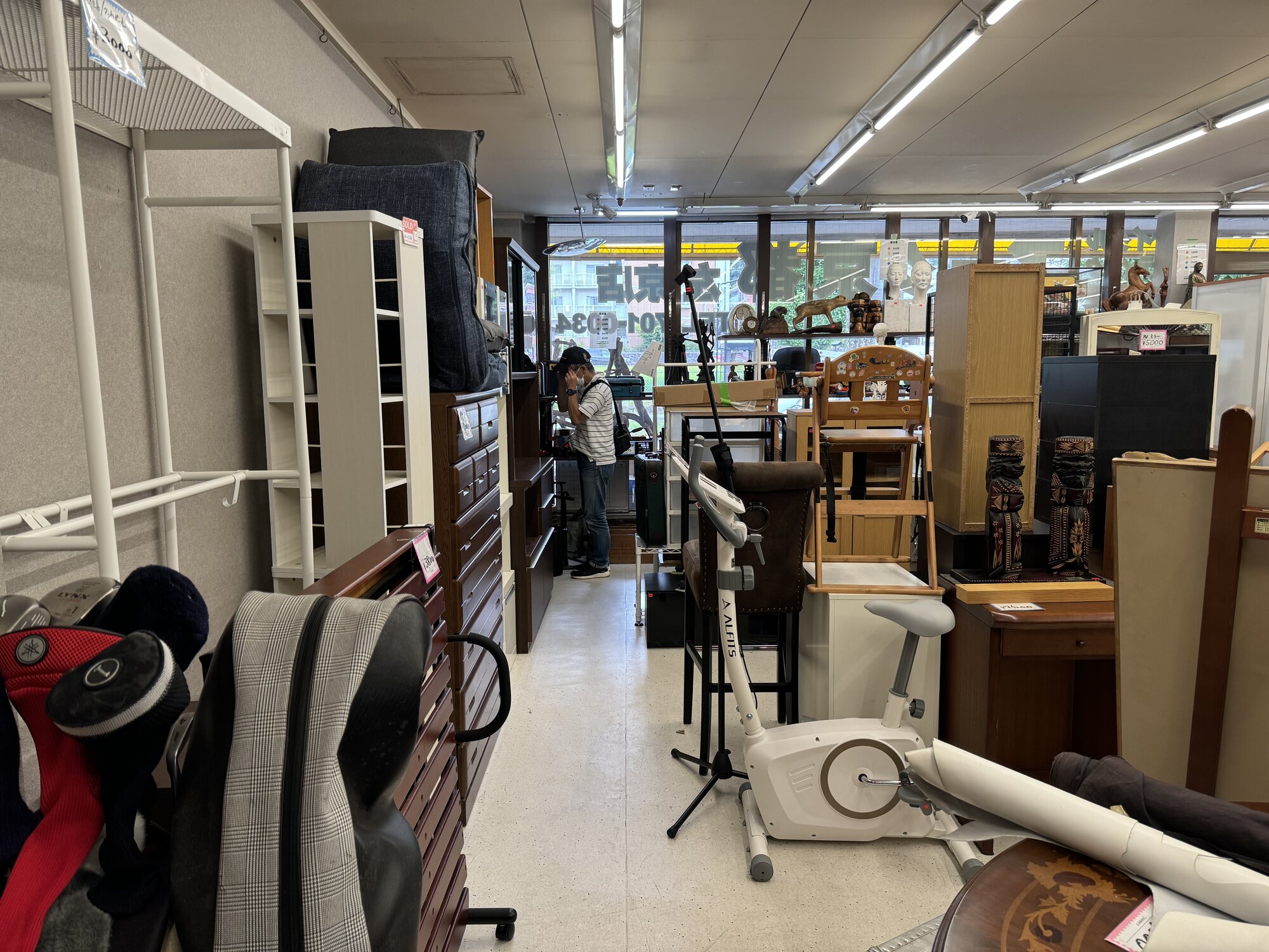 A packed aisle inside a local Kyoto recycling shop, crammed with used furniture, chests of drawers, chairs, golf clubs, an exercise bike and carved wooden folk items under fluorescent light.