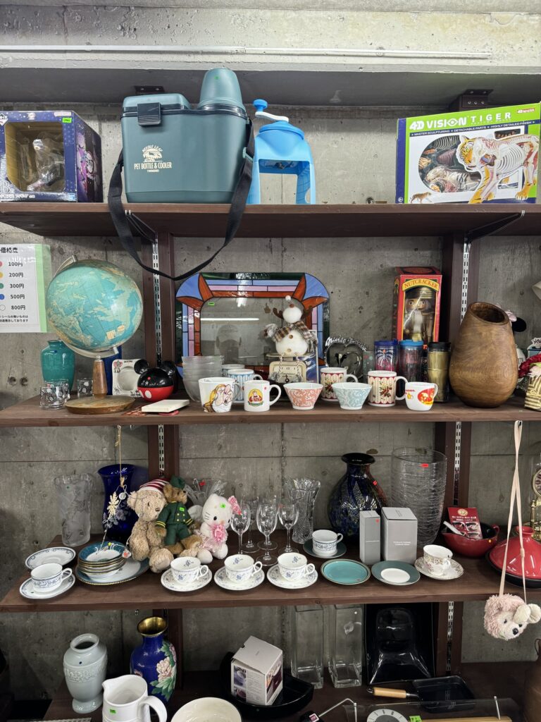 Shelves of secondhand bric-a-brac at a local Kyoto recycling shop: a globe, teacups and mugs, glassware, small vases and figurines packed together on concrete-walled shelving.