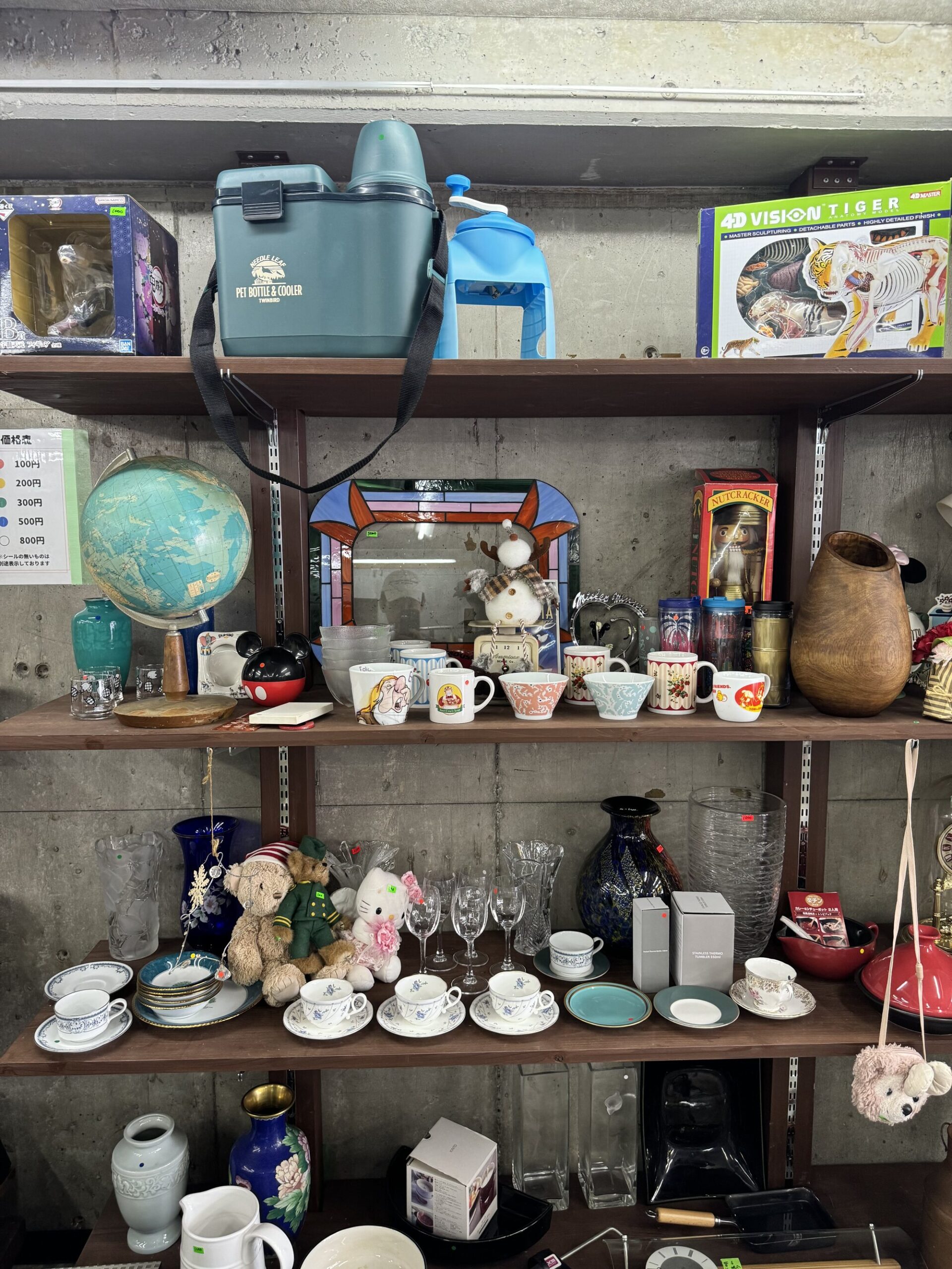 Shelves of secondhand bric-a-brac at a local Kyoto recycling shop: a globe, teacups and mugs, glassware, small vases and figurines packed together on concrete-walled shelving.