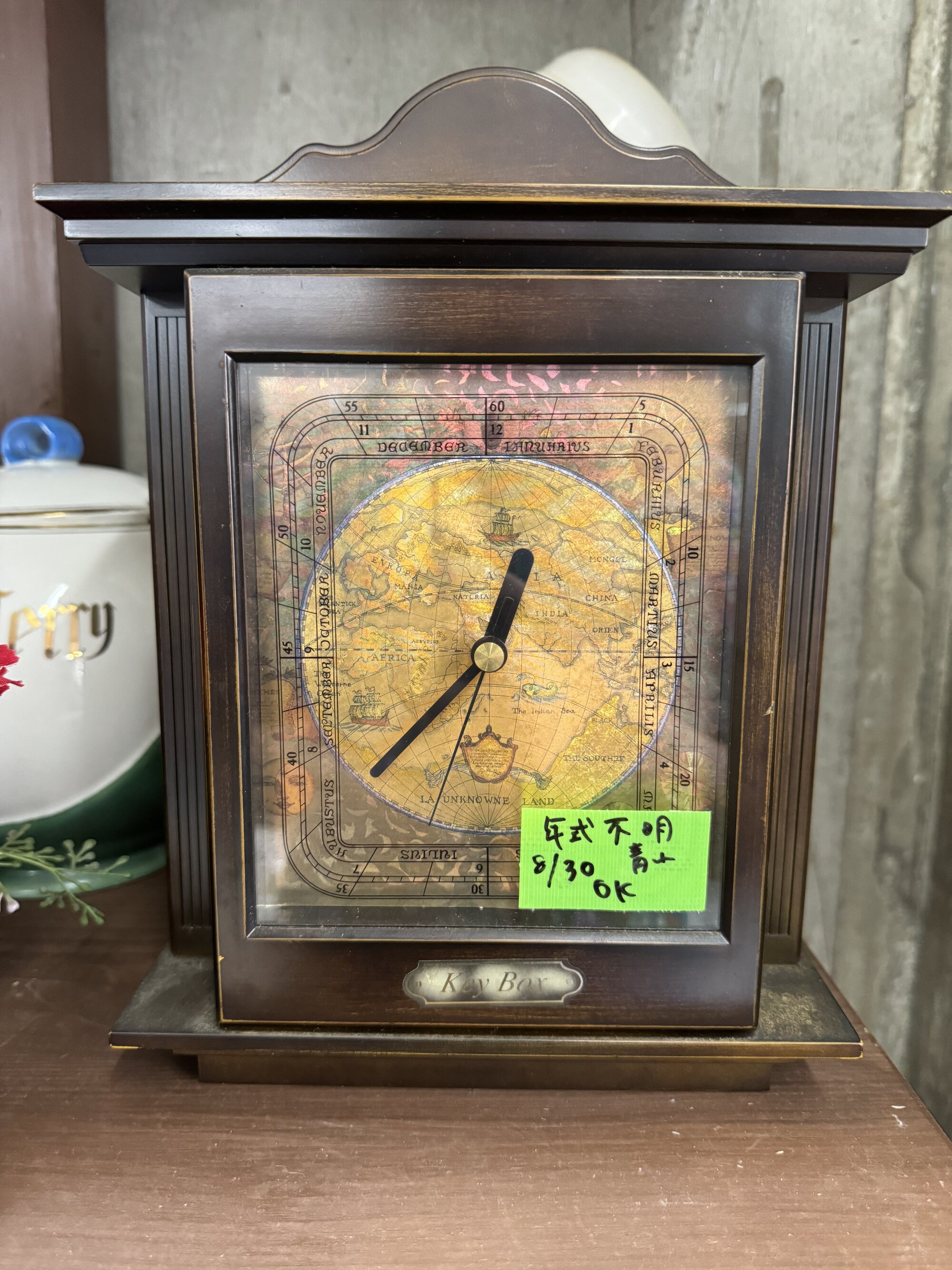 A vintage wooden mantel clock with an antique map-style face and a handwritten style-unknown price tag, on a shelf at a local Kyoto secondhand recycling shop.