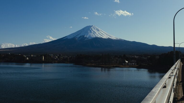 Snow-capped Mount Fuji rising over the calm blue water of Lake Kawaguchi, photographed from the Kawaguchiko Ohashi bridge with the lakeside town of Funatsu along the far shore