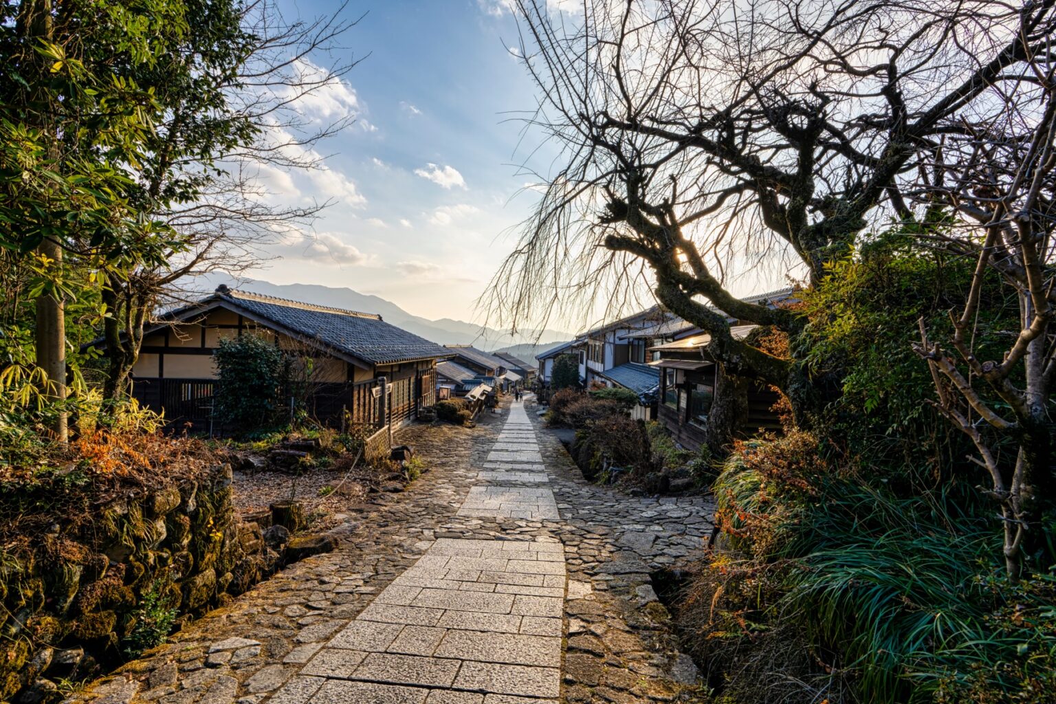 A stone-paved path descending through Magome-juku toward layered blue mountains at sunset with bare trees and traditional Kiso houses lining the slope in winter