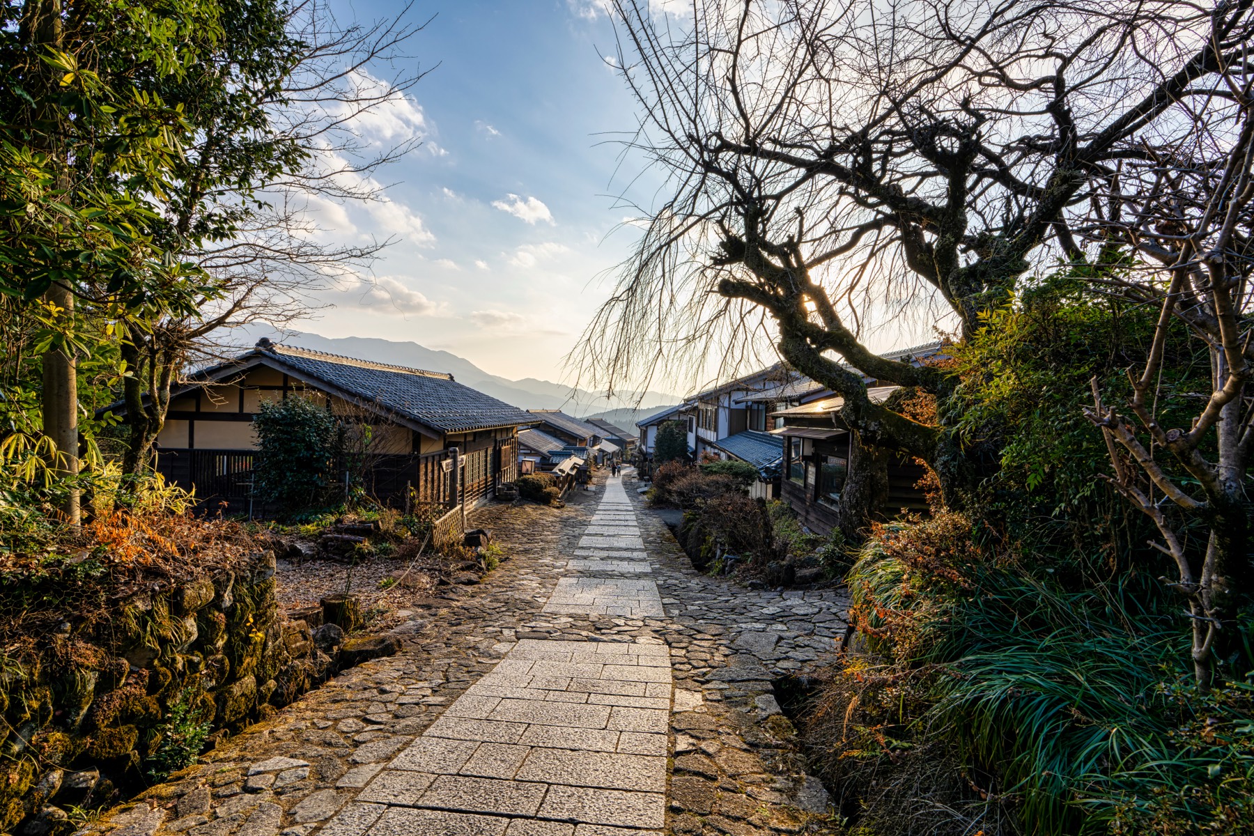 A stone-paved path descending through Magome-juku toward layered blue mountains at sunset with bare trees and traditional Kiso houses lining the slope in winter