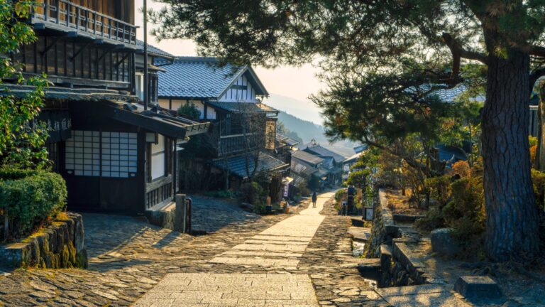 The stone-paved main street of Magome-juku post town curving downhill toward the Kiso mountains and distant Mt Ena at golden hour, lined with dark timber post-town houses and a pine