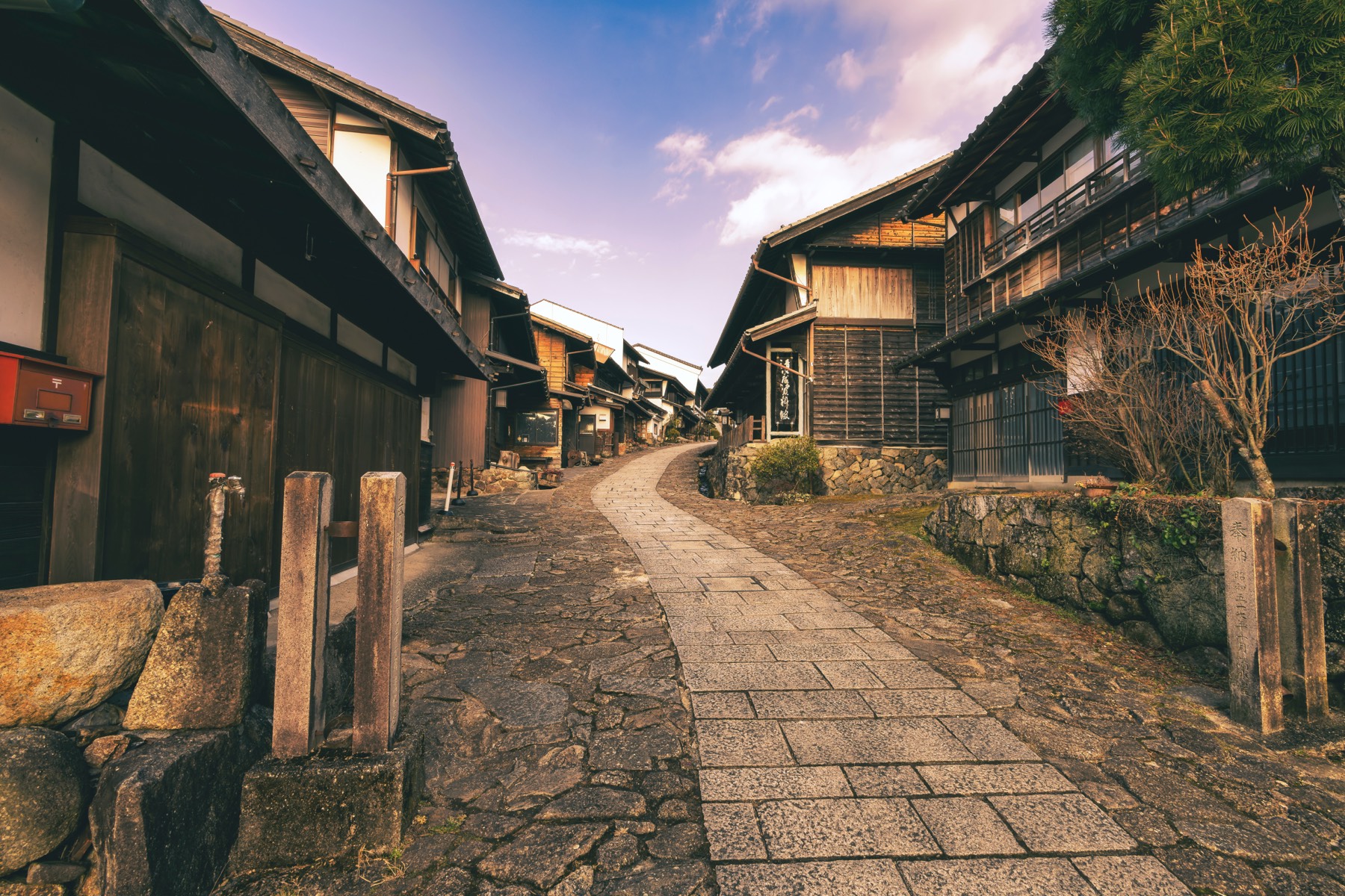 A curving stone-paved street through Magome-juku lined with wooden Edo-period post-town buildings and an old red Japanese post box on the left in soft evening light