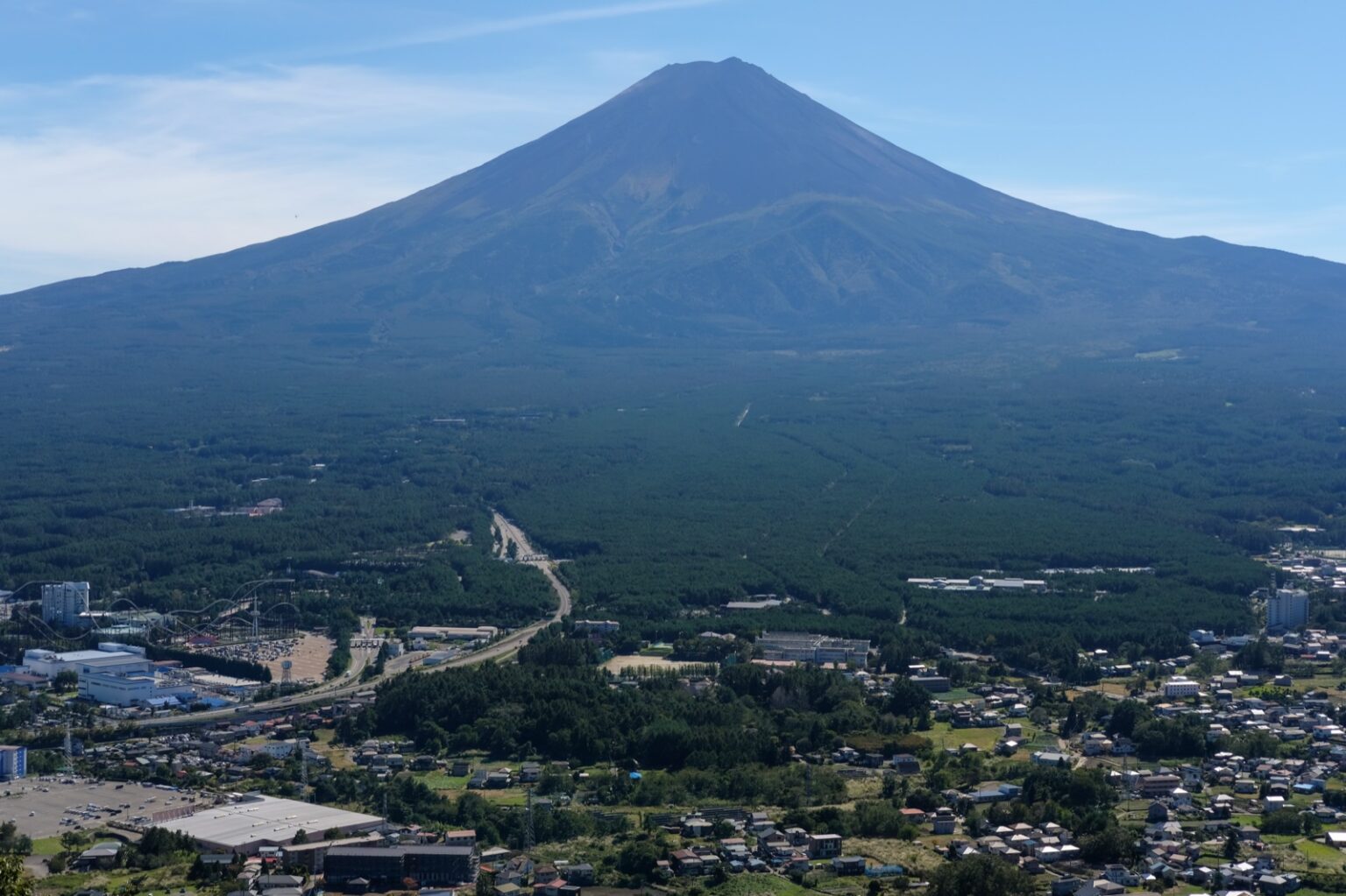 Mt Fuji rising above the Fuji Five Lakes basin, with the town and an amusement park spread across the forest below, seen from the Kawaguchiko panorama viewpoint