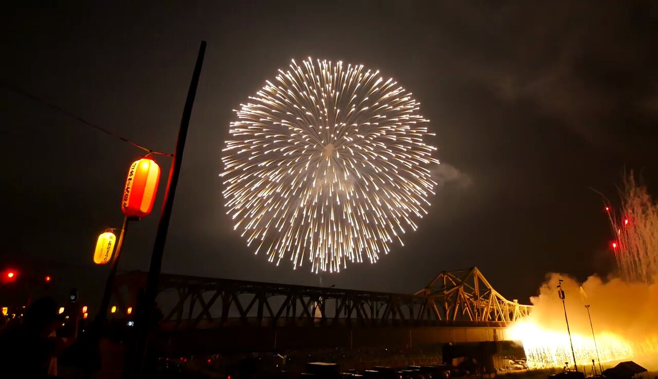 A single sanjakudama 90-centimetre firework shell bursting over the Shinano River railway bridge at the Nagaoka fireworks festival, with red paper lanterns in the foreground