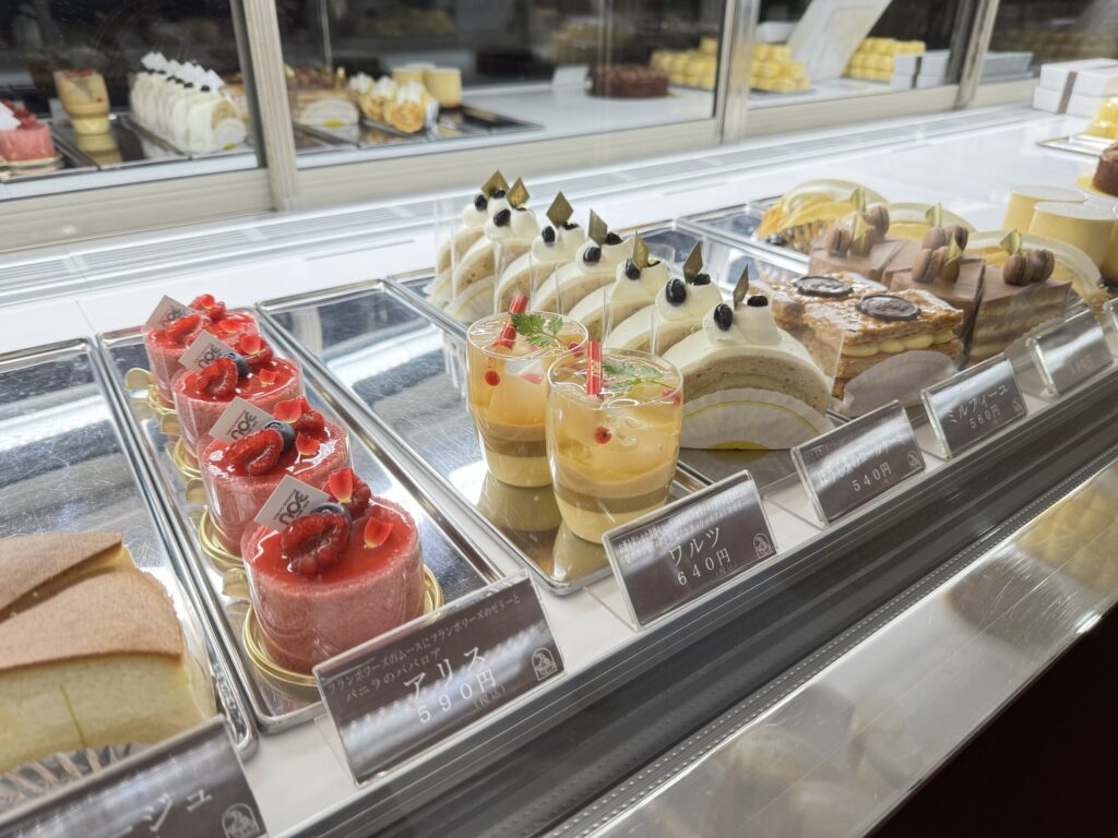 A glass patisserie case at Okashinoie Noe in Nerima, Tokyo, lined with strawberry shortcakes, cream roll cakes and cup desserts, each hand-labelled, in the shop's warm-lit front counter.