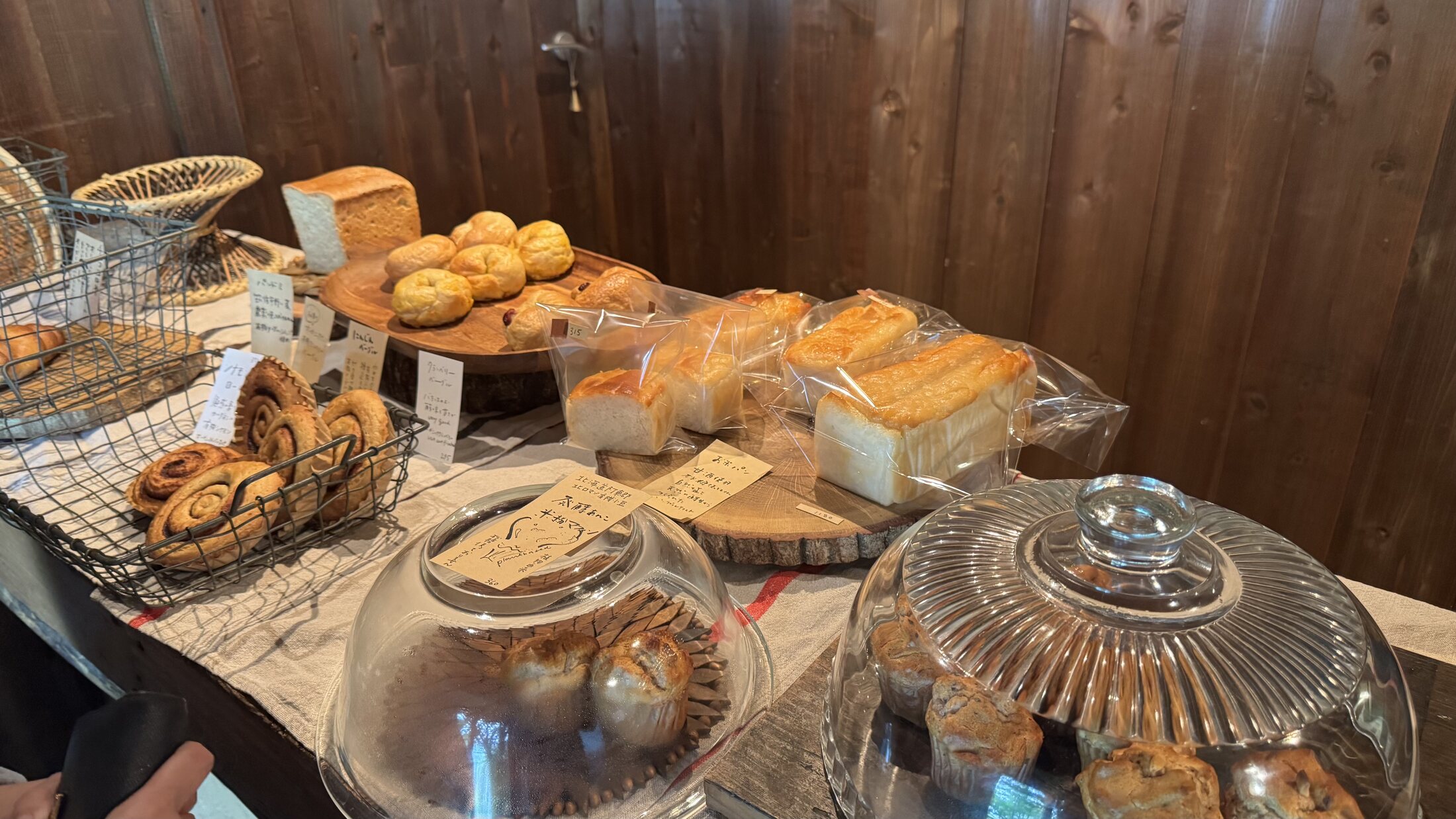 The bread display at Pandian bakery in Fujikawaguchiko: baskets and glass domes of natural-yeast loaves, buns and muffins with handwritten labels on a rustic counter.